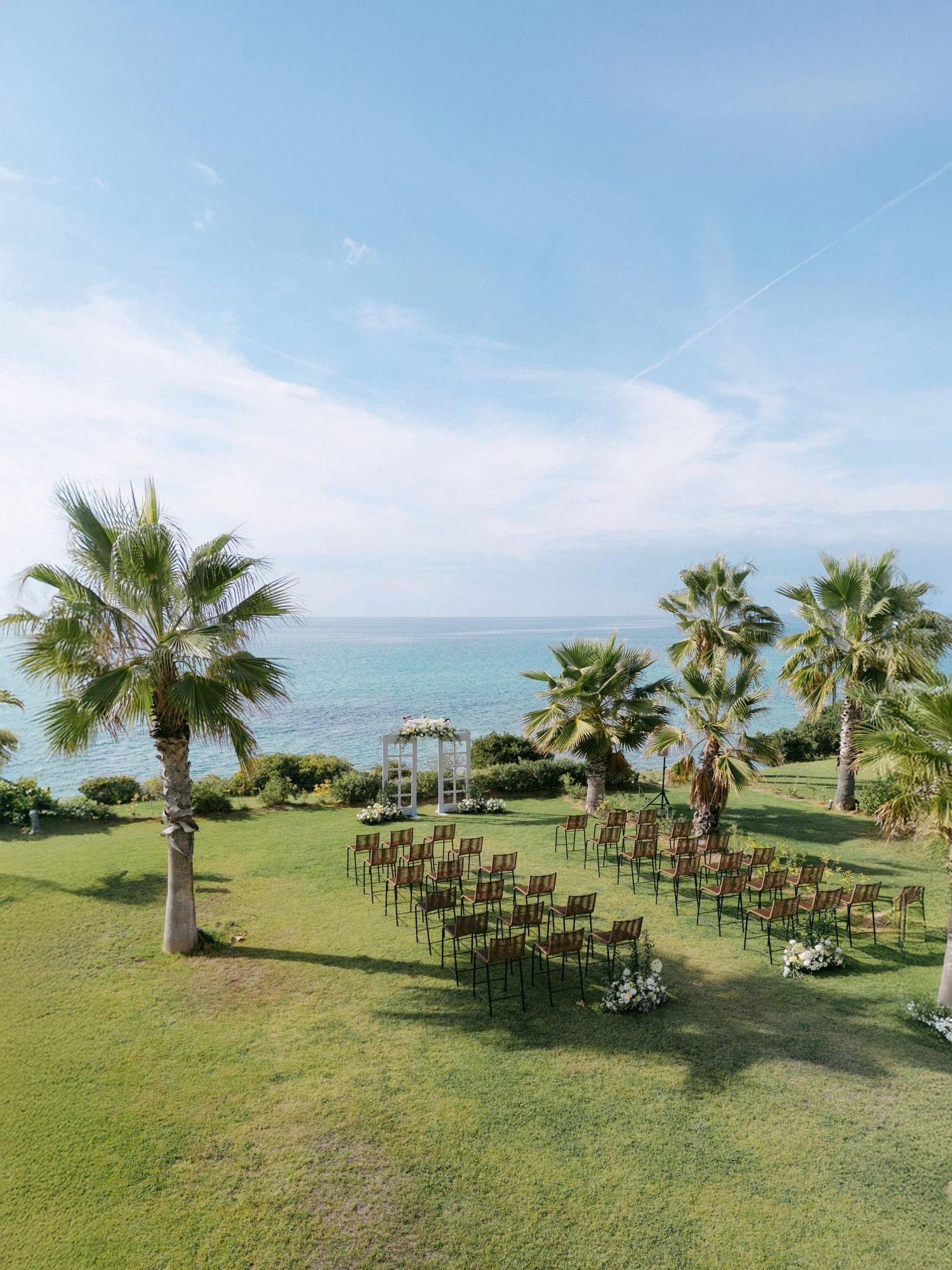 Outdoor wedding ceremony setup with rows of chairs on a green lawn overlooking the ocean with palm trees framing the scene.