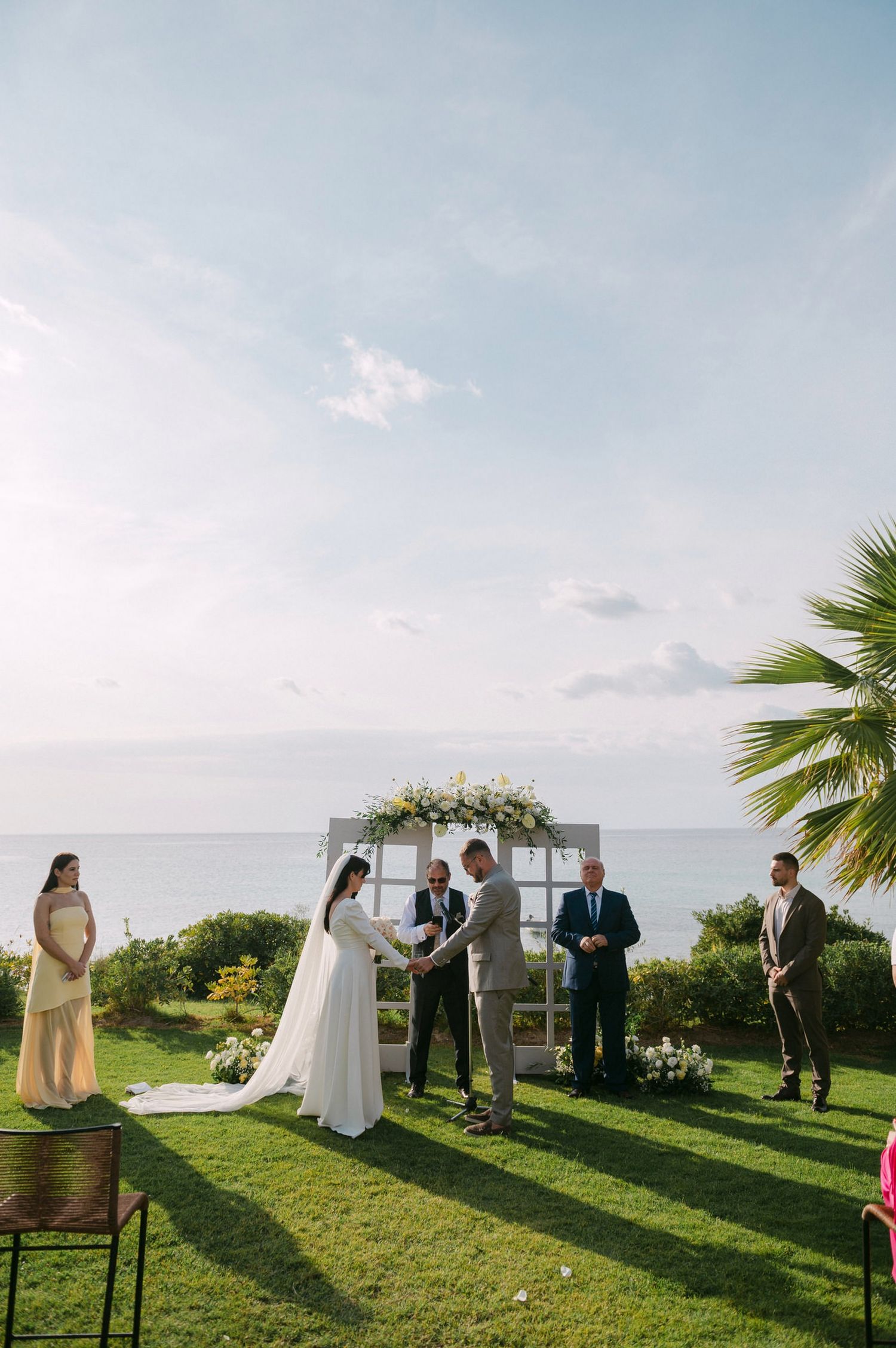 An outdoor wedding ceremony takes place on a grassy cliff overlooking the ocean with palm trees and a floral arch at sunset.