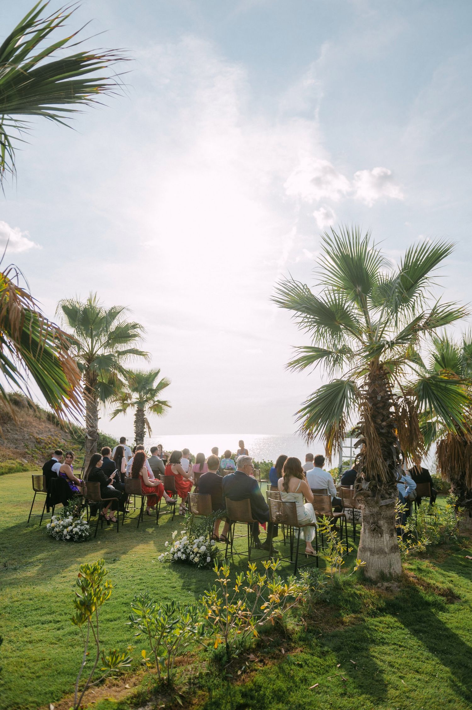 Guests gather under palm trees at an outdoor wedding ceremony on a grassy hillside overlooking the ocean at sunset.
