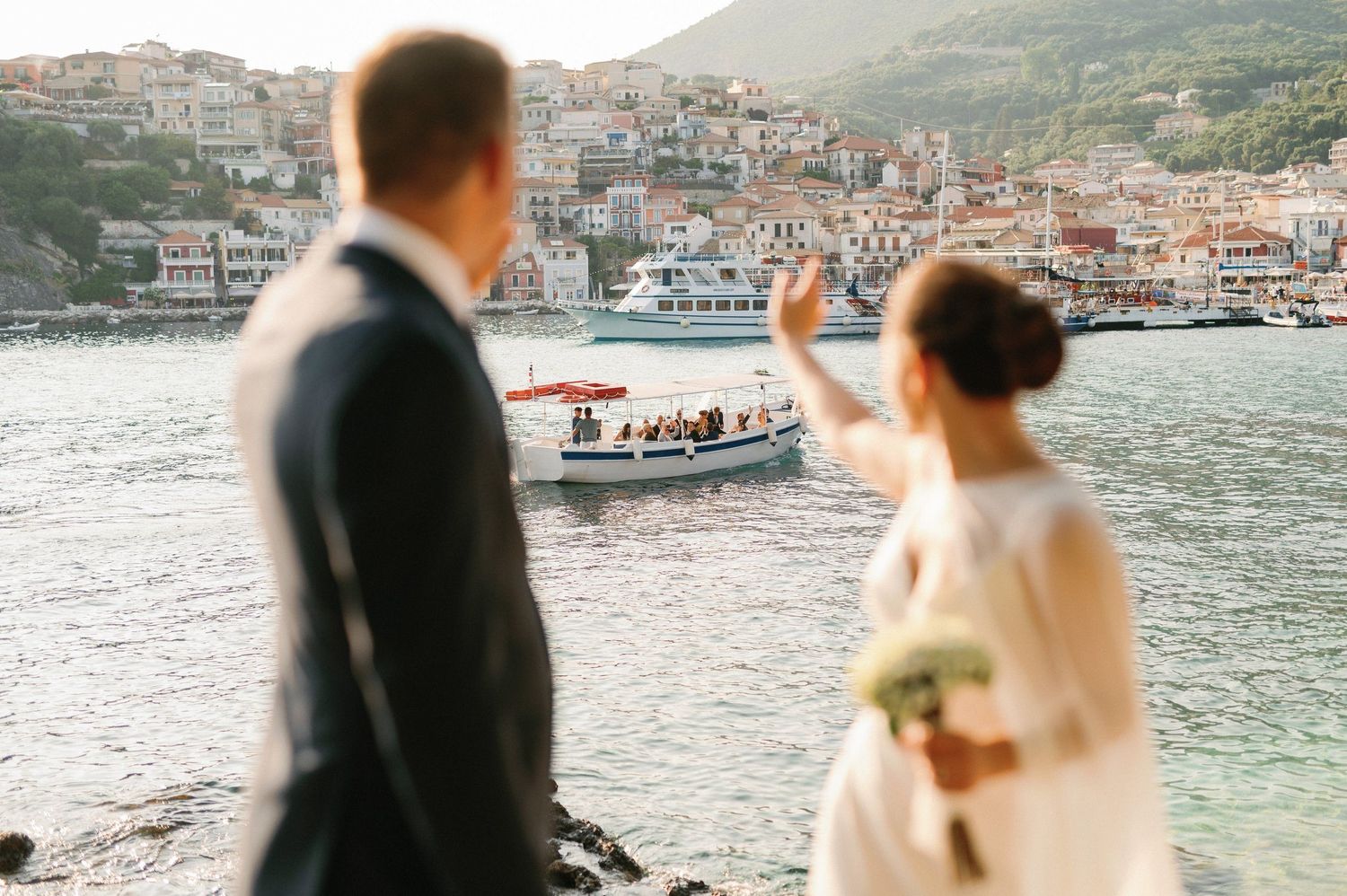Couple gazes at scenic coastal town view with boats in harbor and hillside buildings along Mediterranean waterfront at Parga Greece.