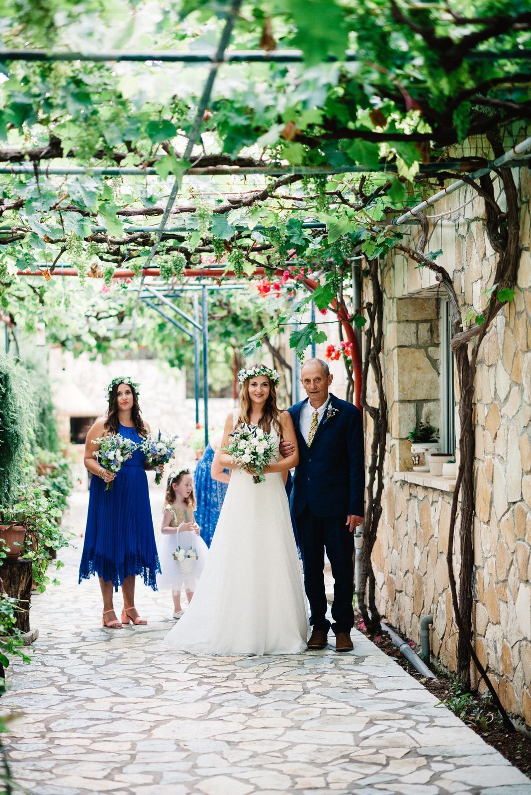 Wedding ceremony under a vine-covered stone archway with bride in white and attendant in blue.