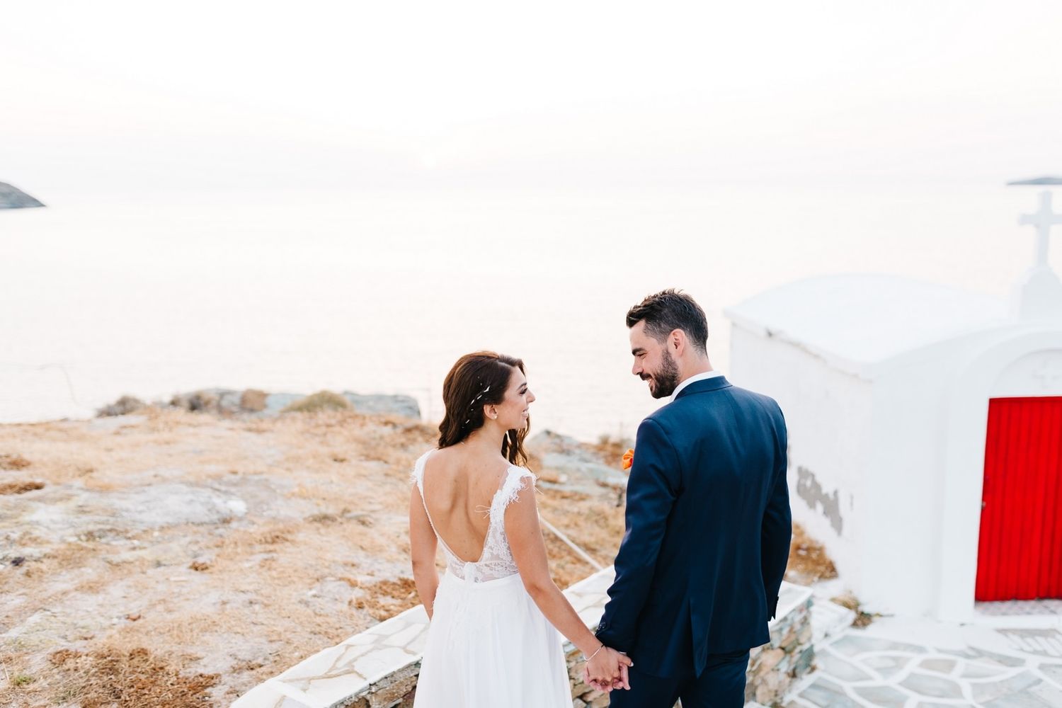Wedding couple holding hands near white building with red door overlooking ocean.