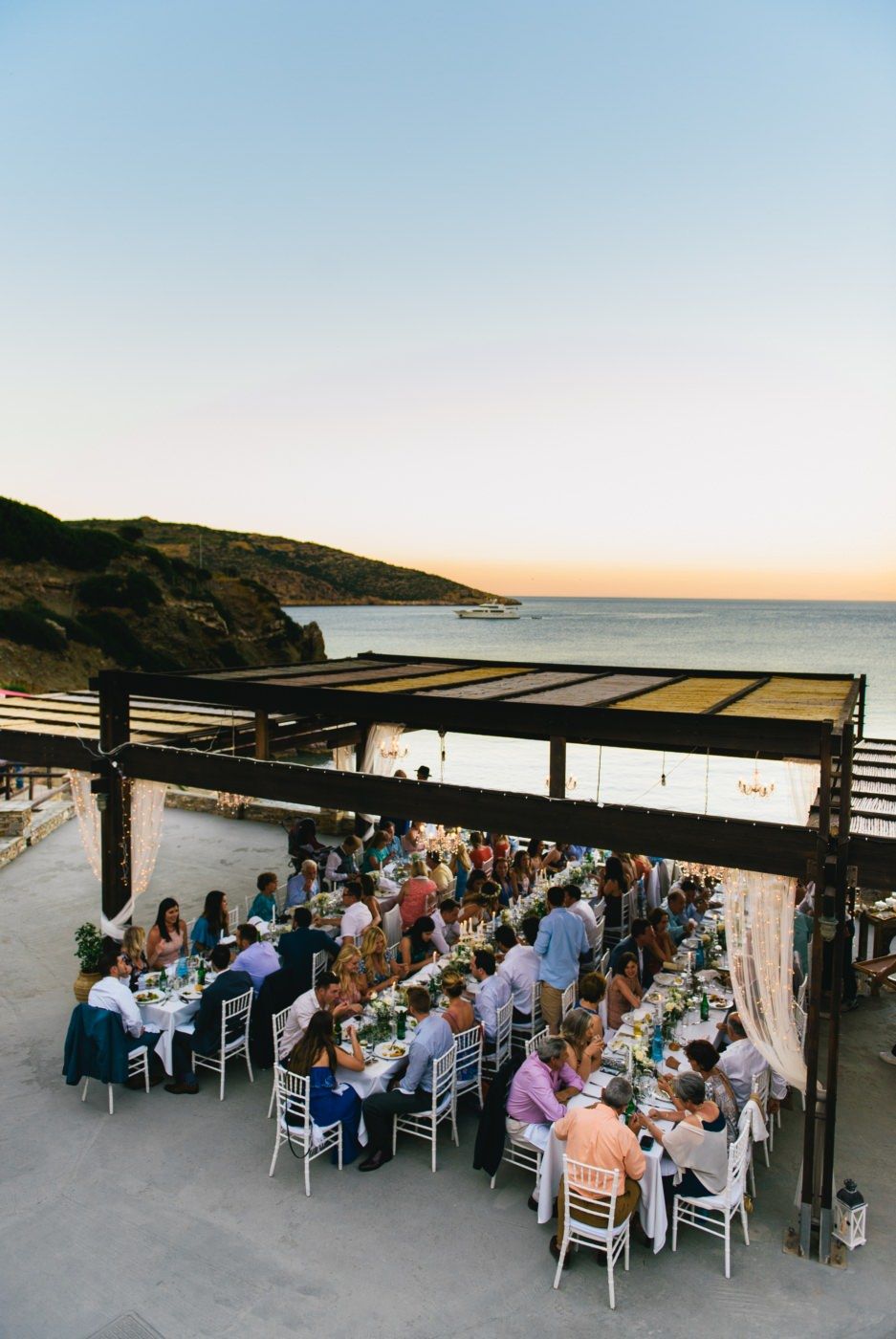 Outdoor wedding reception under covered terrace by the ocean at sunset with long dining tables.
