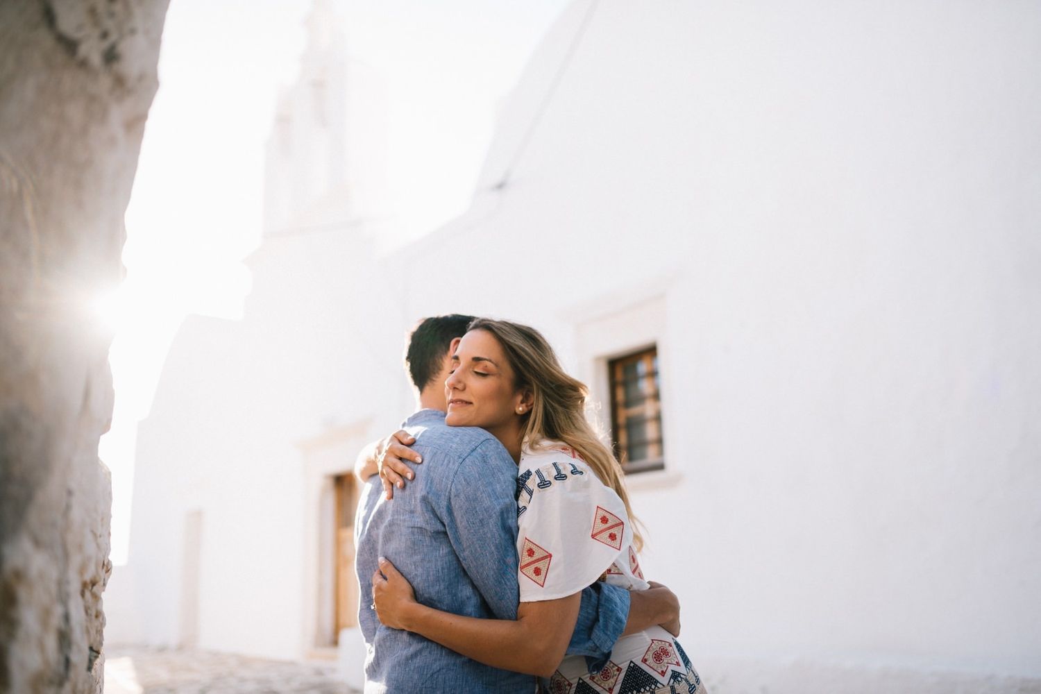 A tender embrace against a white Mediterranean building in soft sunlight.