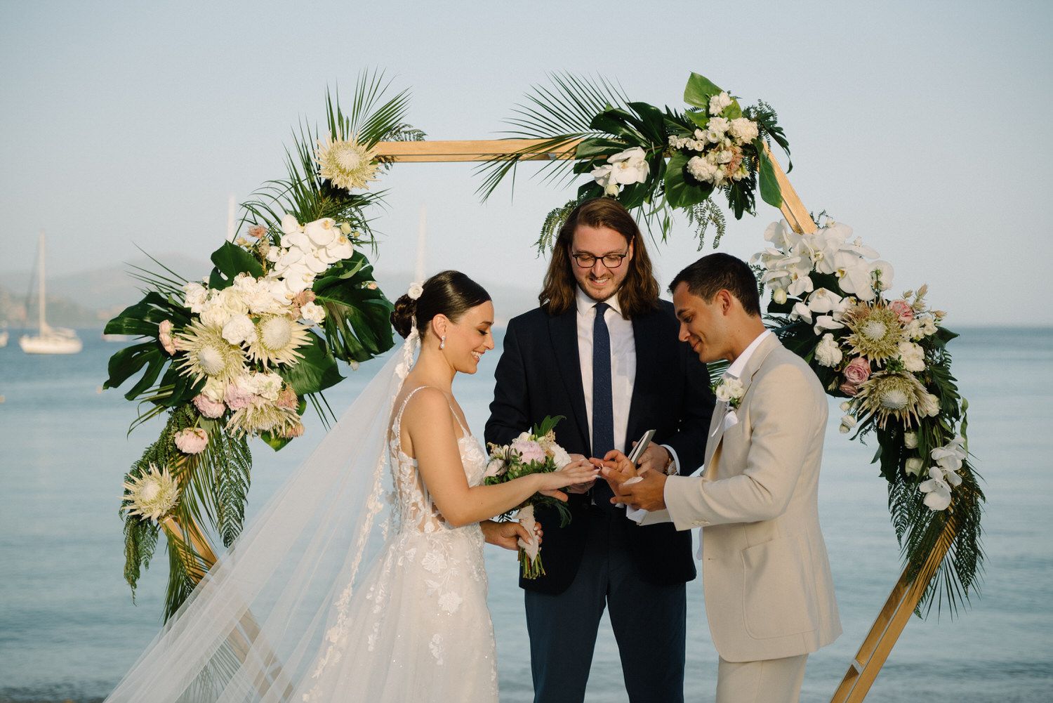 Wedding couple exchanging rings under floral arch at waterfront ceremony.
