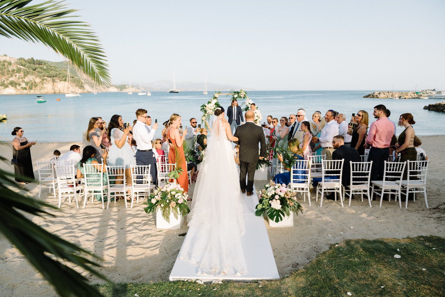 Beachfront wedding ceremony with white chairs and tropical floral arrangements.