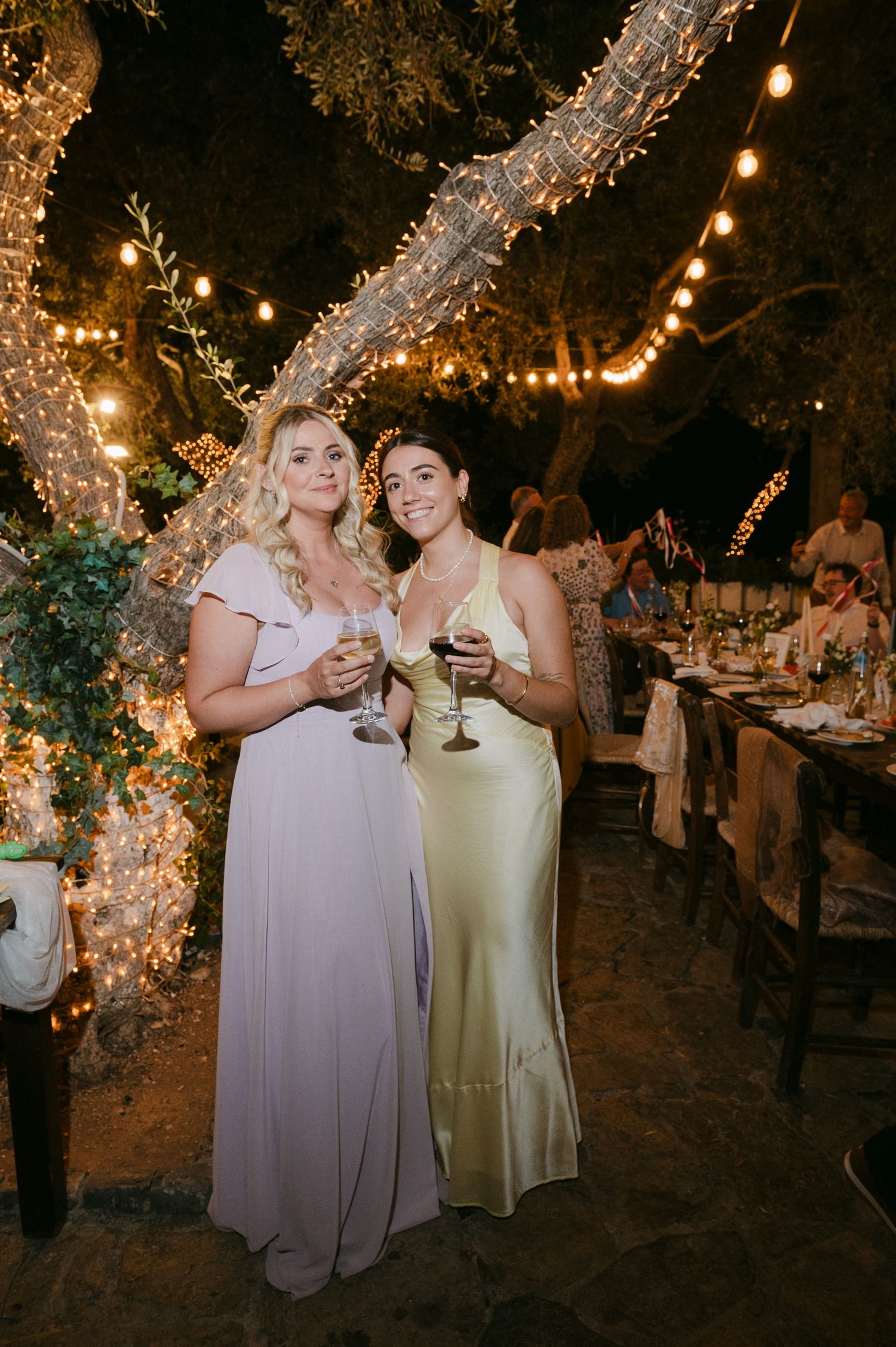 Two guests in elegant dresses enjoy drinks at an outdoor evening reception with string lights and decorative branches.