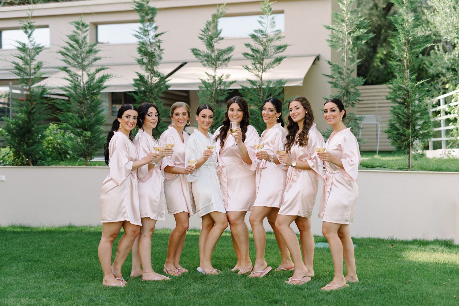 A bridal party in matching pink robes poses together on a grassy lawn.