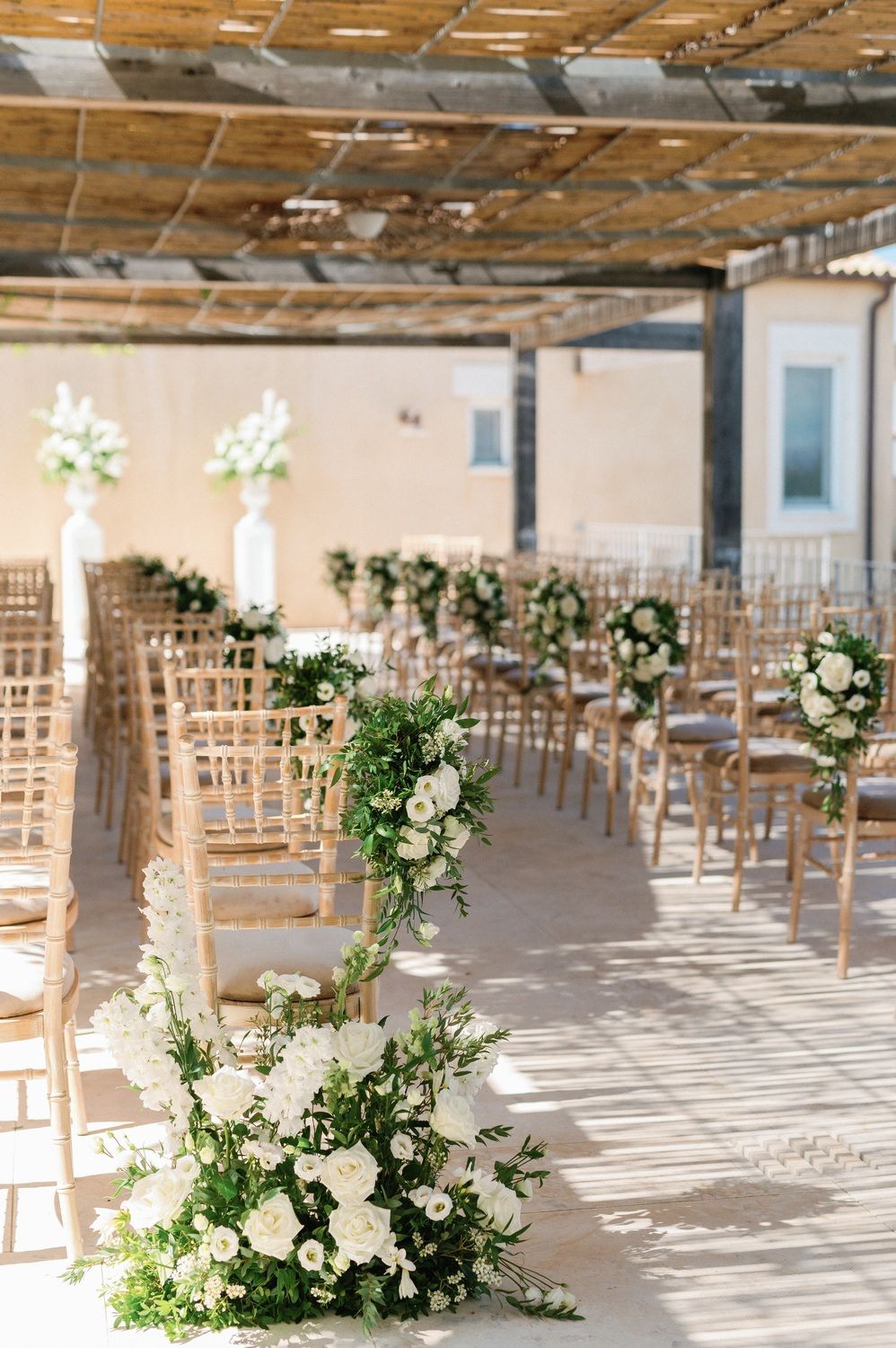 Wedding ceremony venue with elegant white floral arrangements and wooden chairs under a rustic bamboo ceiling.