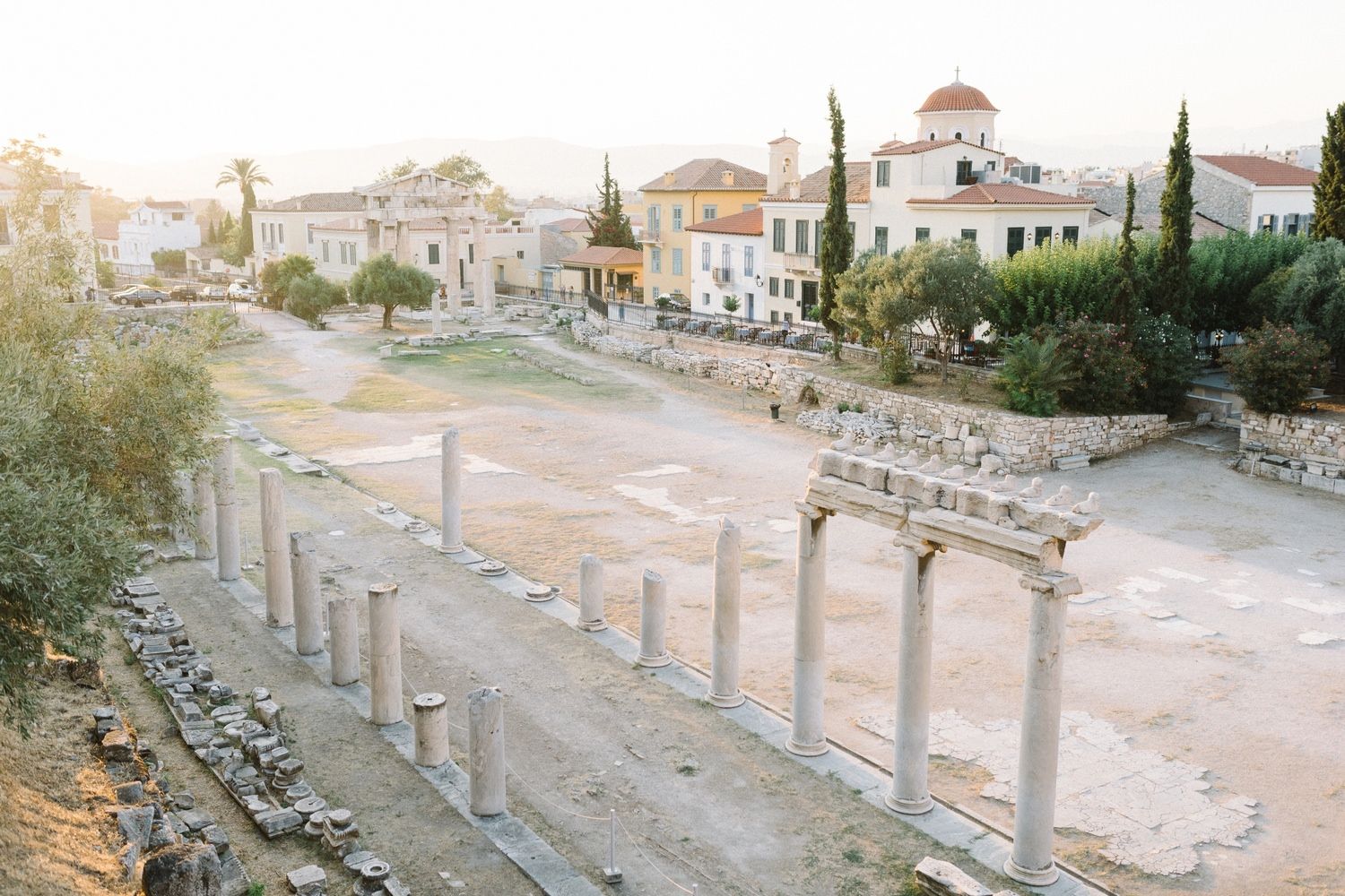 Ancient stone columns line the street in Greece with historic buildings and cypress trees in the background.