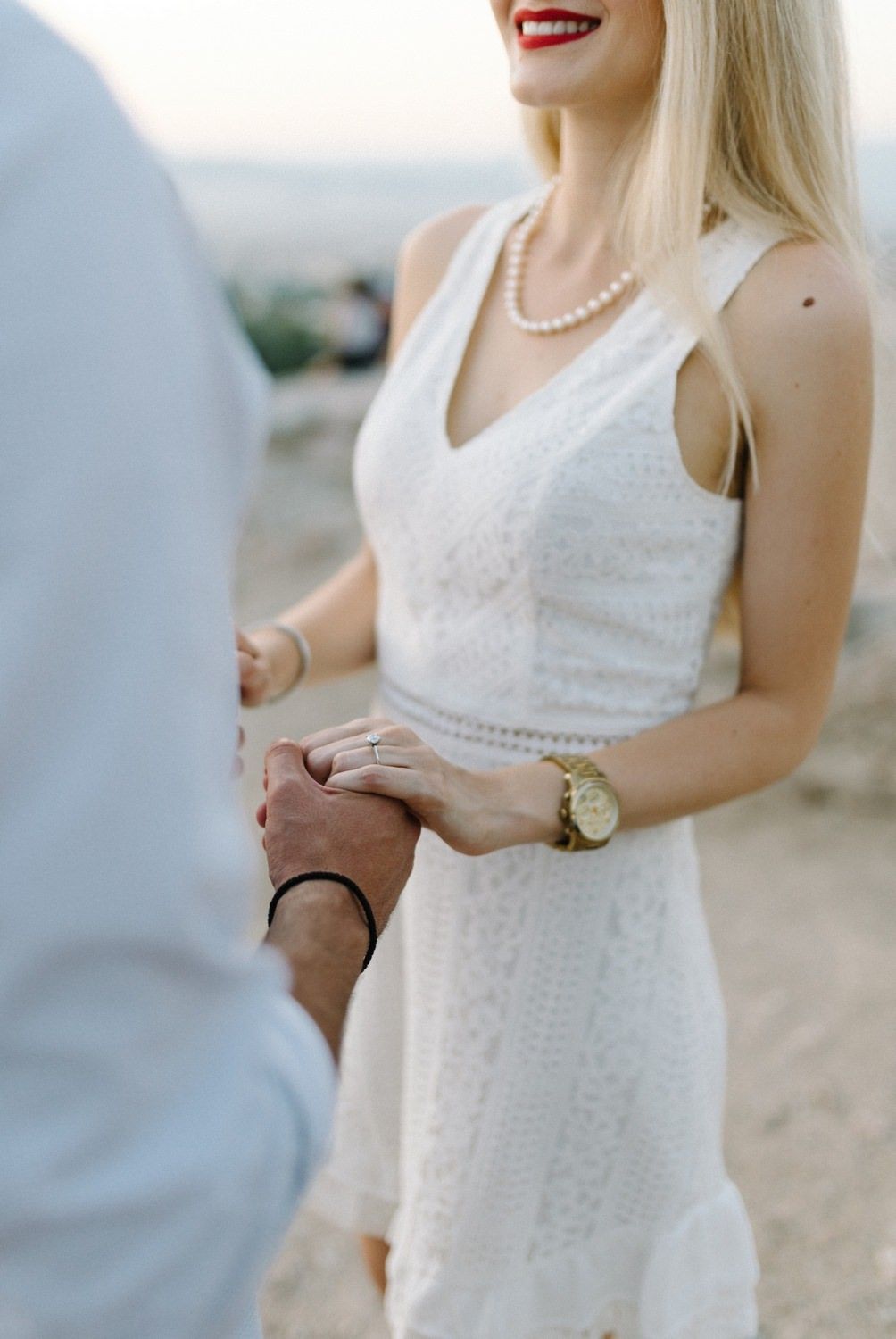 Woman in white dress with pearl necklace and gold watch holding hands with partner.