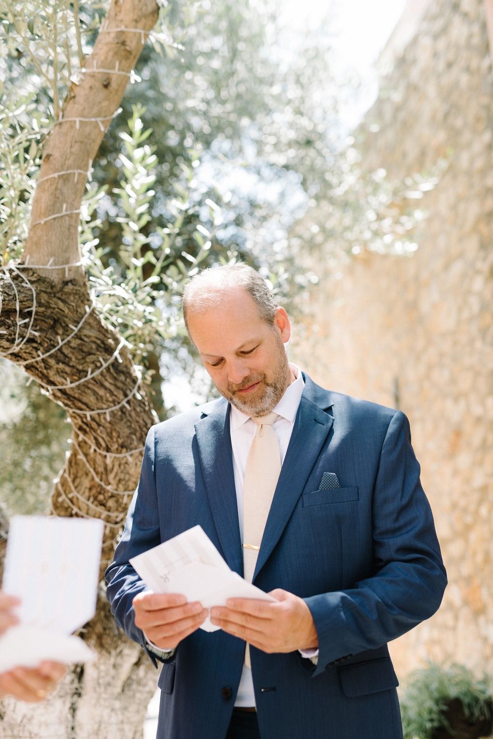 Person in navy suit reading a letter outdoors surrounded by olive trees.