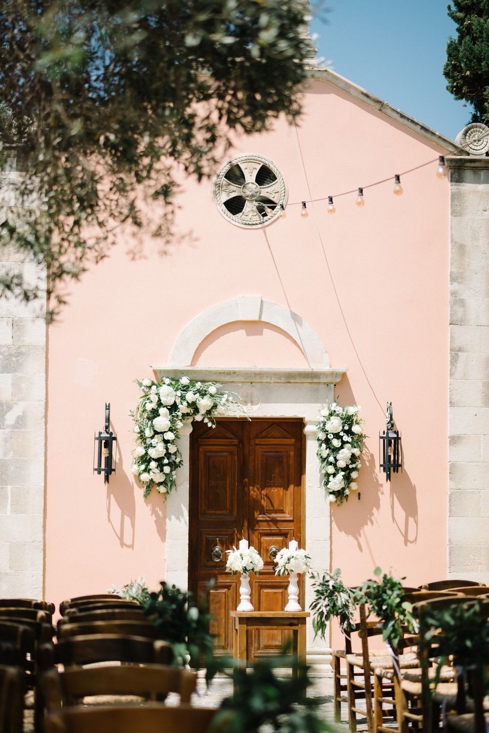 Blush pink chapel entrance decorated with white floral arrangements and wooden chairs.