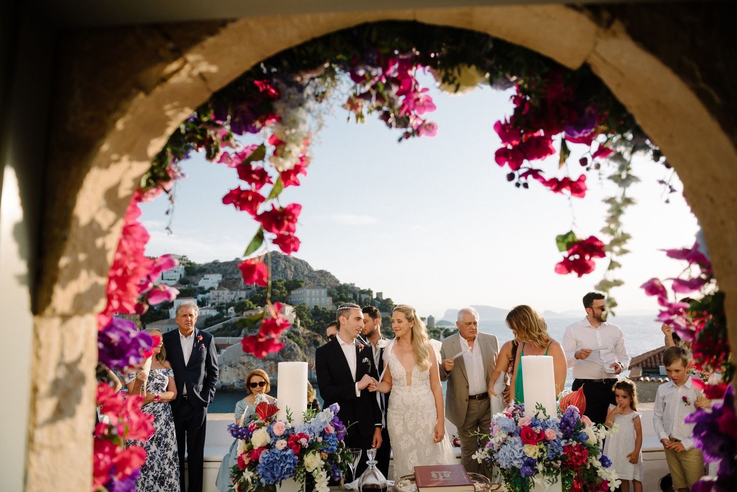 Wedding guests gather under a floral arch with pink bougainvillea and mountain views.