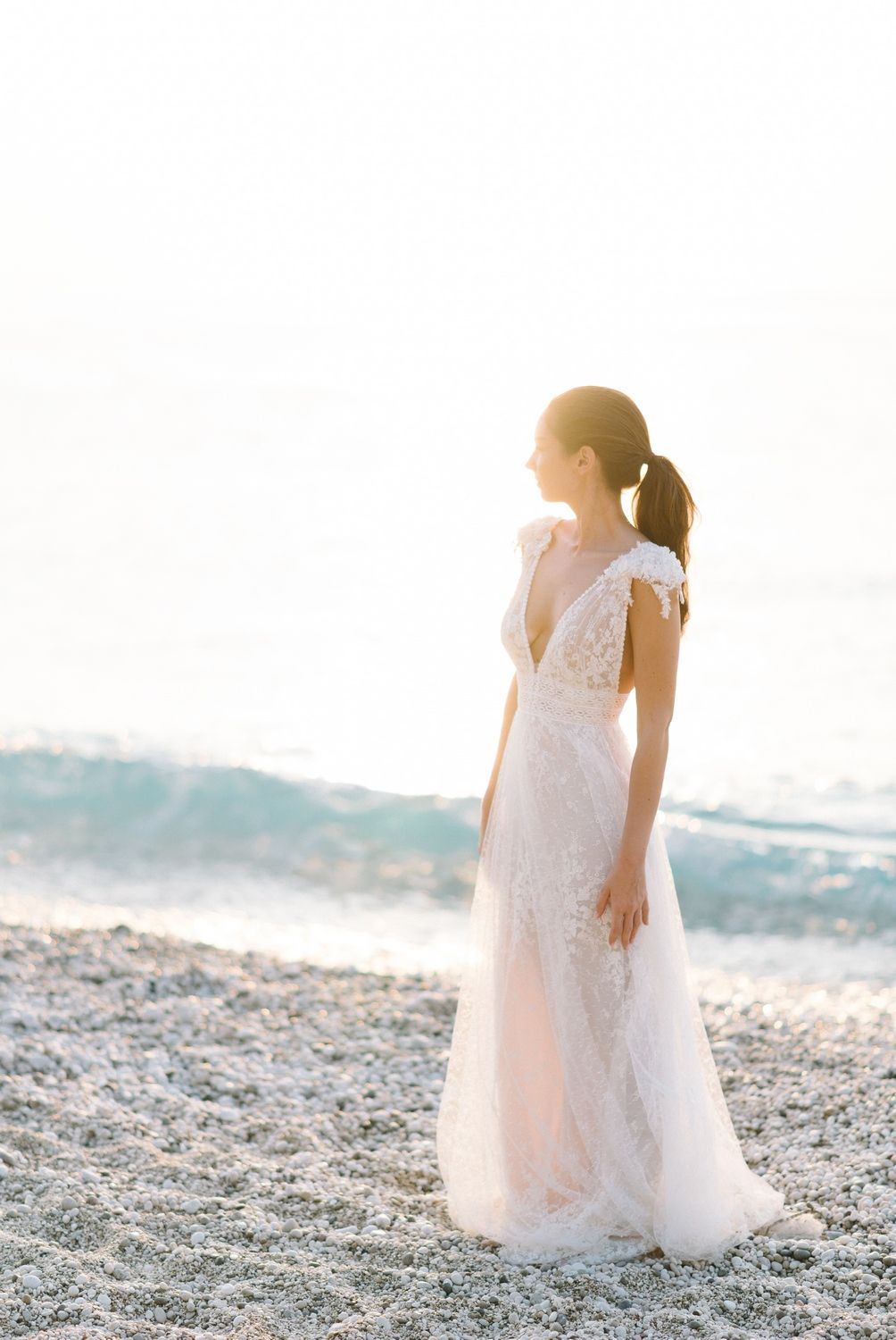 Elegant bride in lace V-neck wedding dress standing alone on sunlit beach.