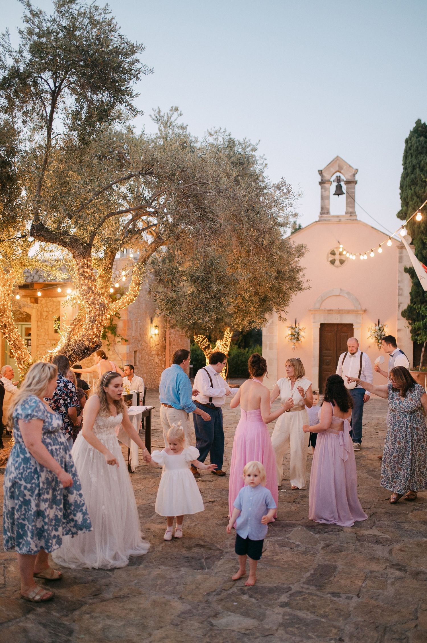 Wedding guests dance and celebrate outdoors at dusk near a mission-style church with string lights and olive trees.
