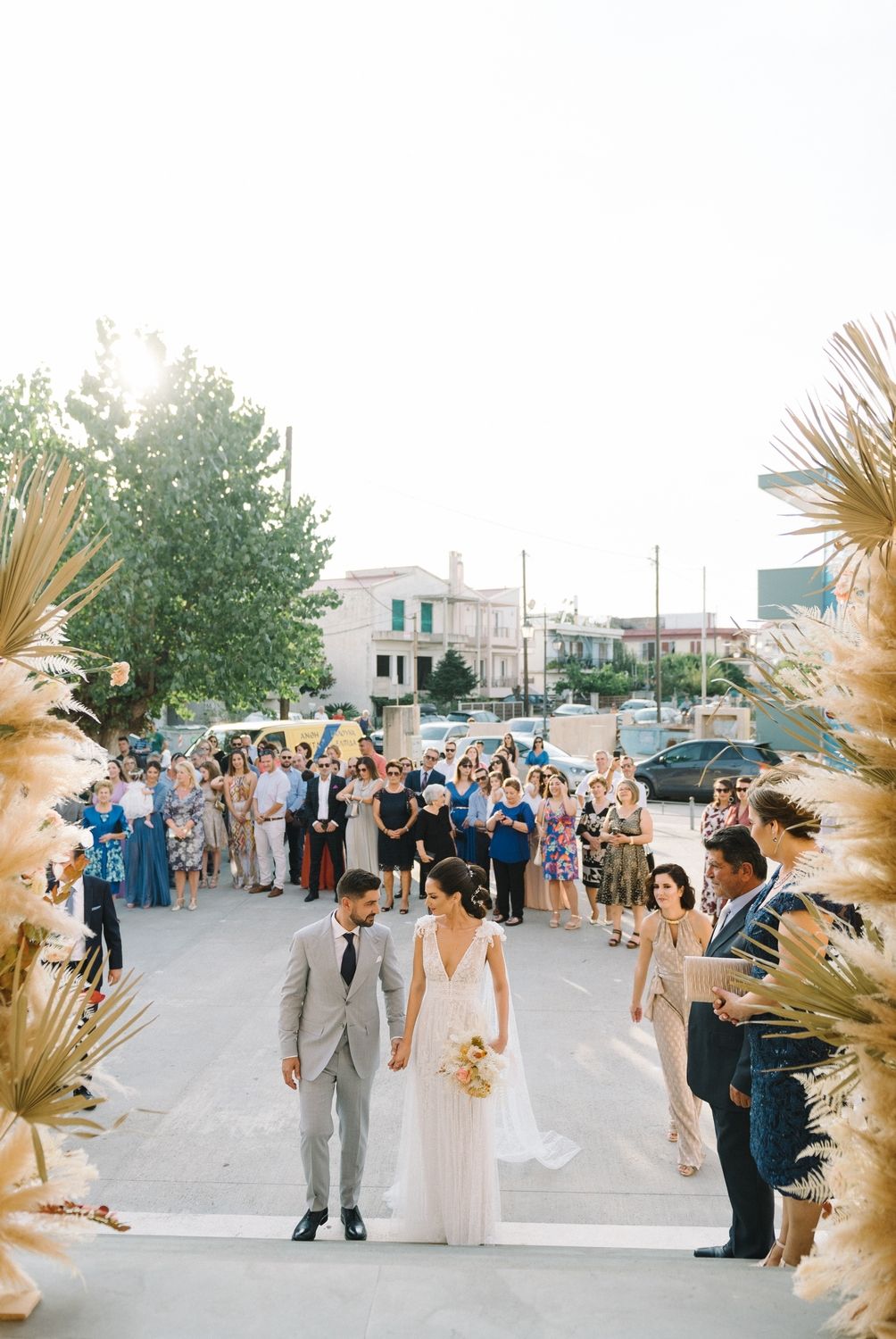 Newlyweds walking through pampas grass arch as guests celebrate on urban street.