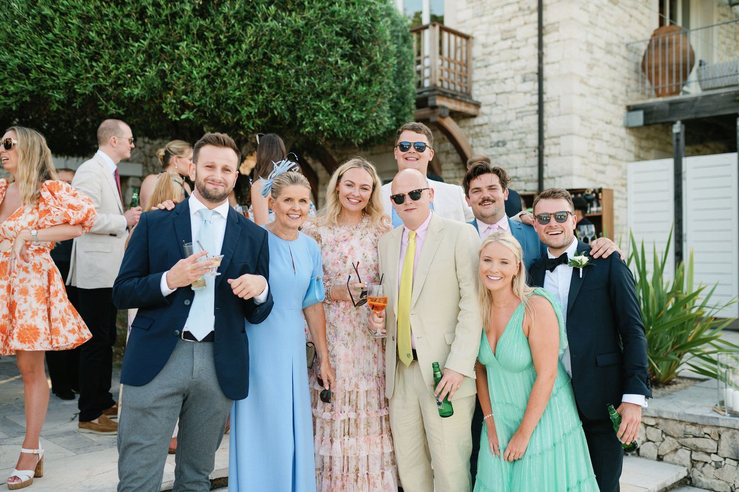 Wedding guests in pastel attire gather for group photo at outdoor venue.