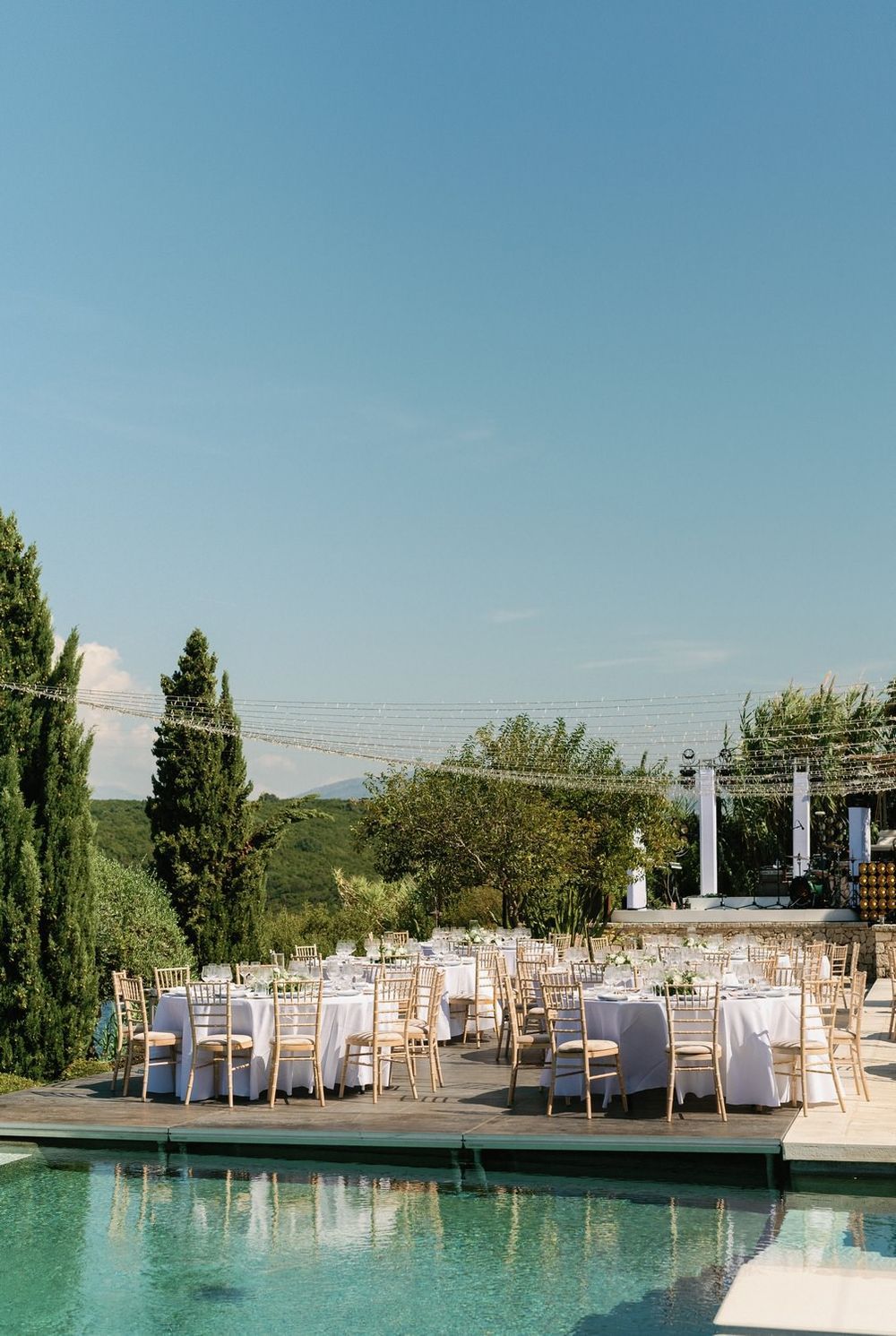Elegant outdoor wedding reception setup with white chairs arranged around a swimming pool under a bright blue sky.