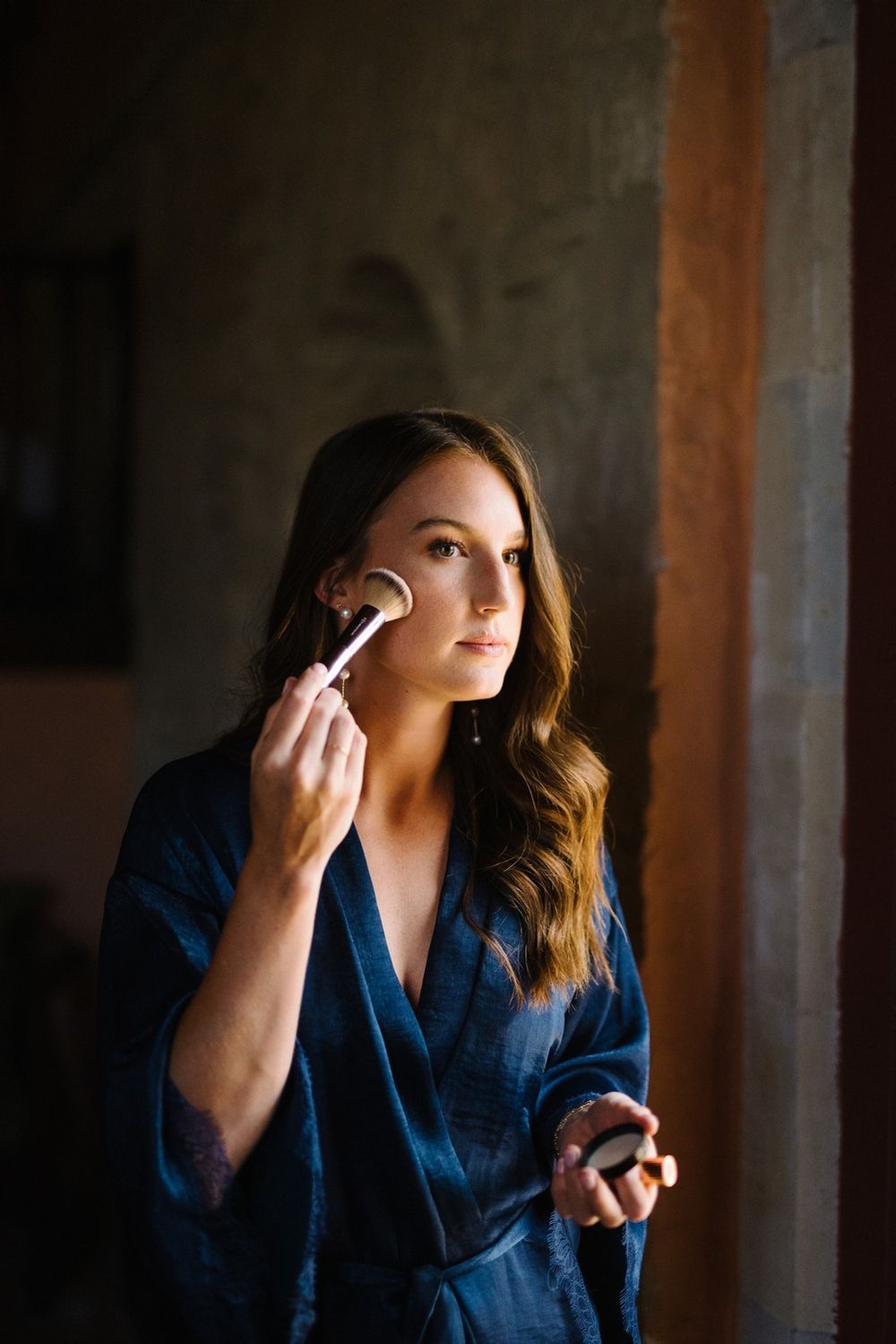 A person applying makeup with a brush while wearing a navy blue robe in moody lighting.
