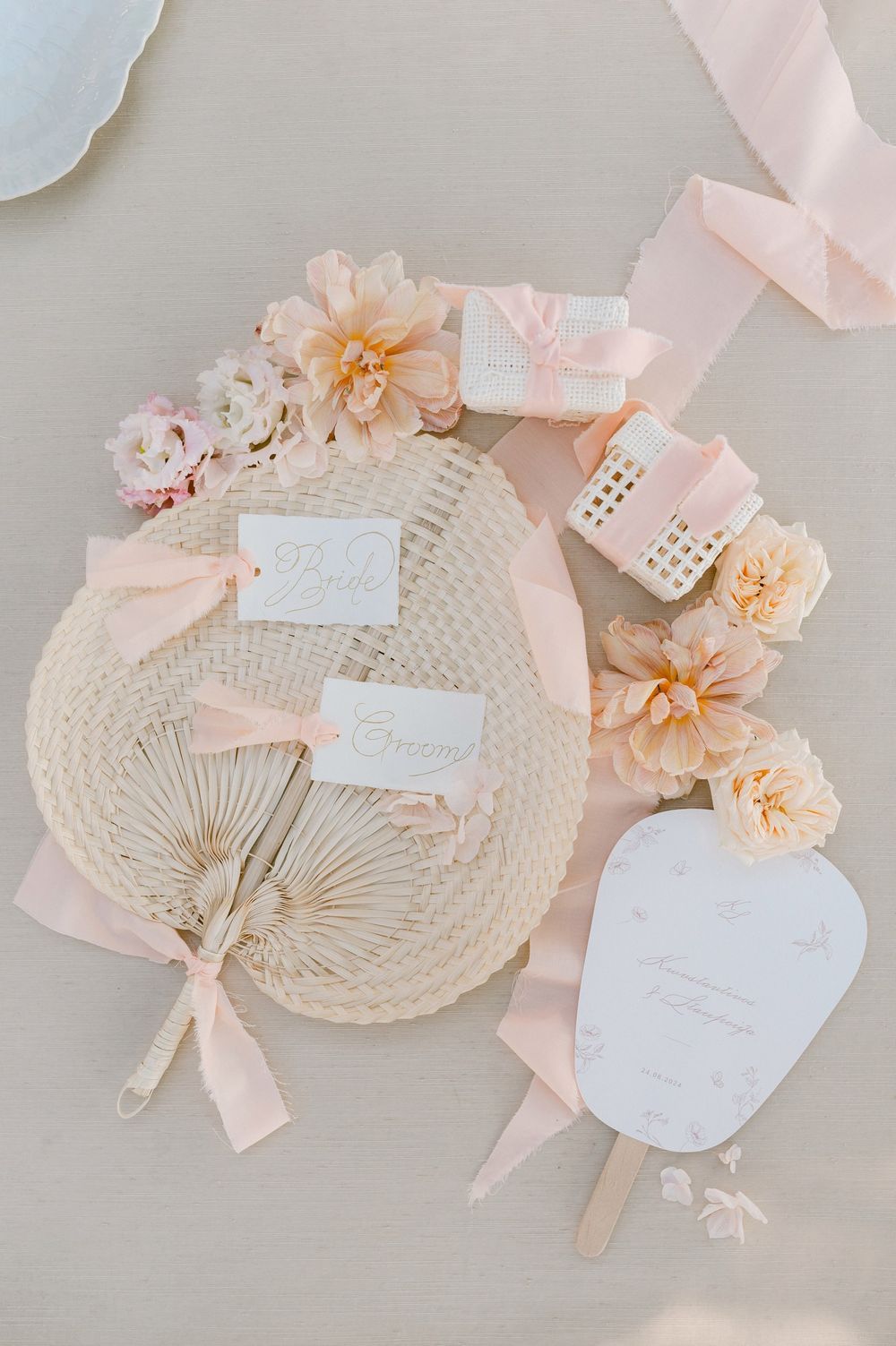 Delicate pink and cream wedding favors with paper fans, silk ribbons, and peach floral accents arranged on light surface.
