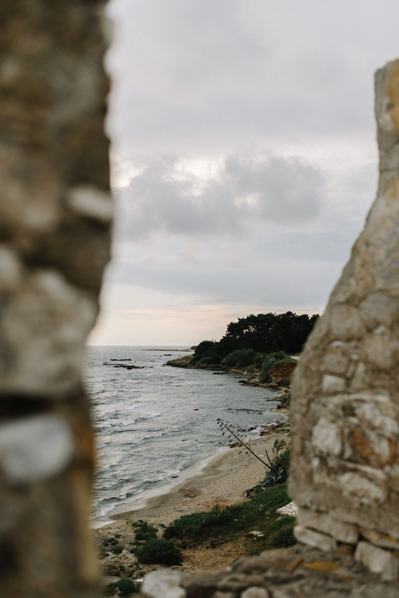 Scenic coastal view through stone ruins showing rocky beach and cloudy sky.
