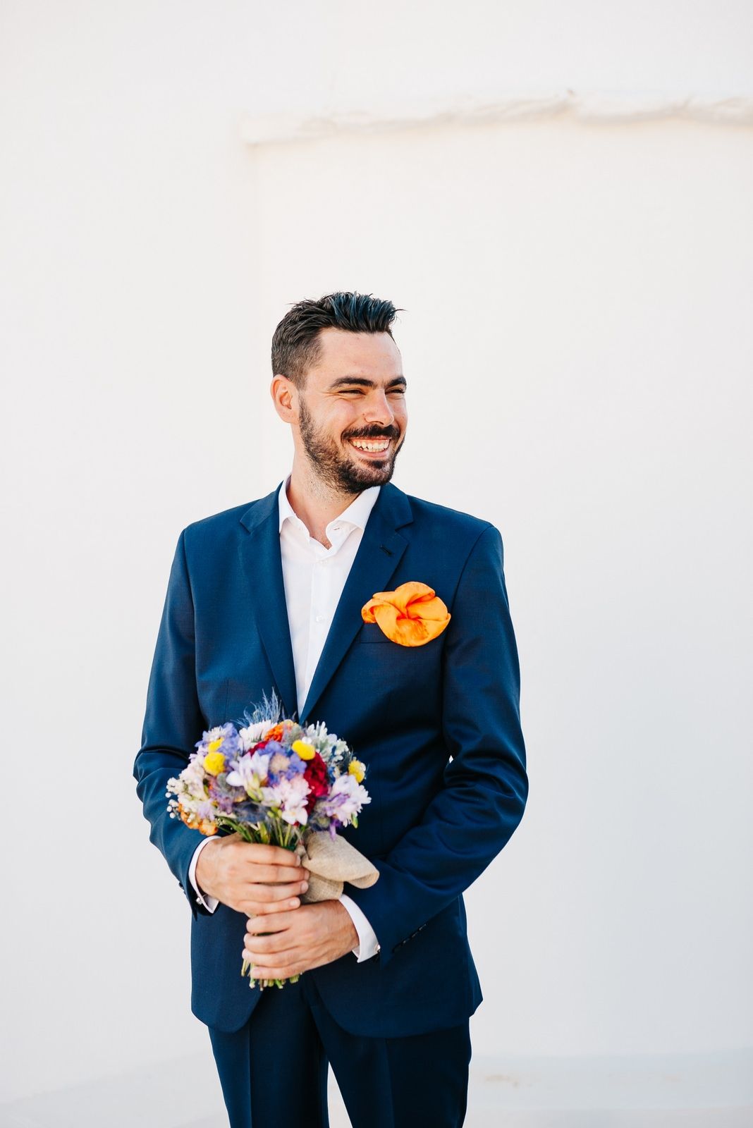 Groom in navy suit holding colorful wedding bouquet against white wall.