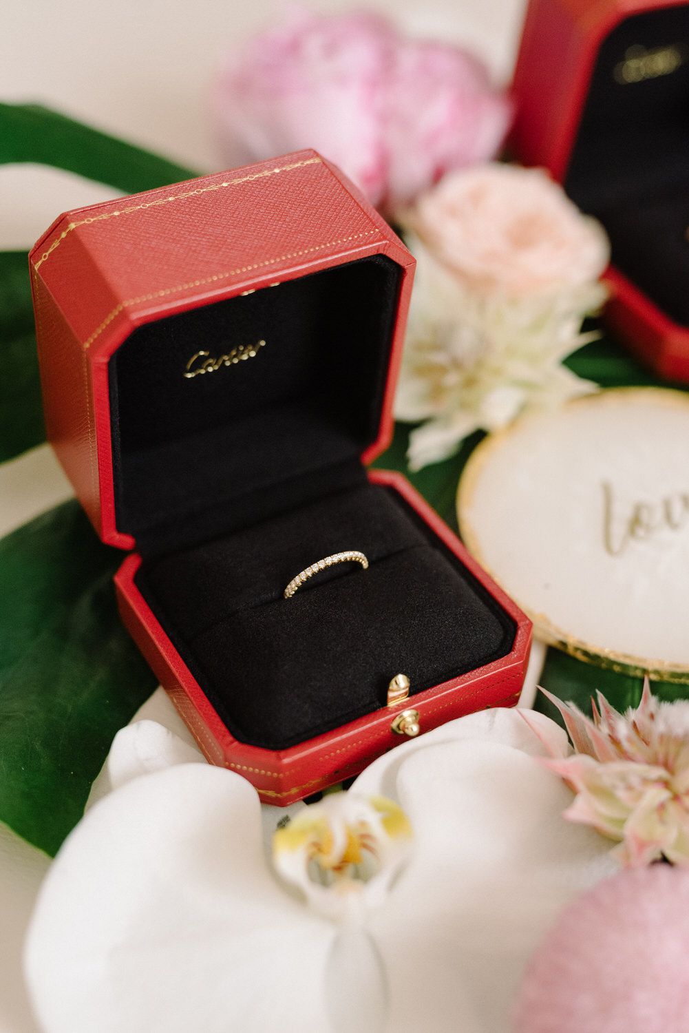 Delicate wedding ring displayed in red Cartier box with flowers and orchids.