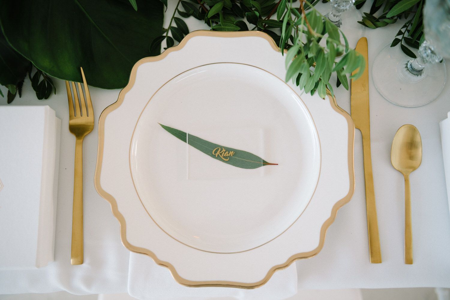 An elegant place setting with white plate, gold flatware, and green leaf accent.
