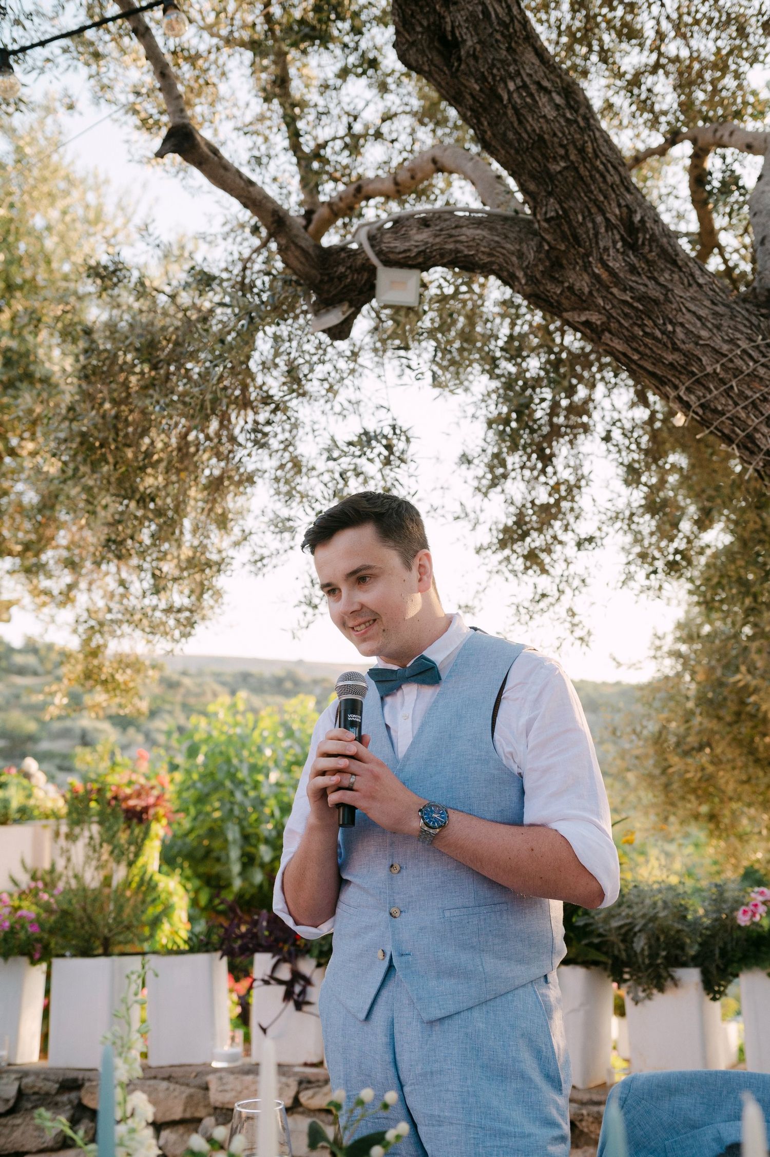 Someone in a light blue vest giving an outdoor speech at a rustic venue beneath oak trees.