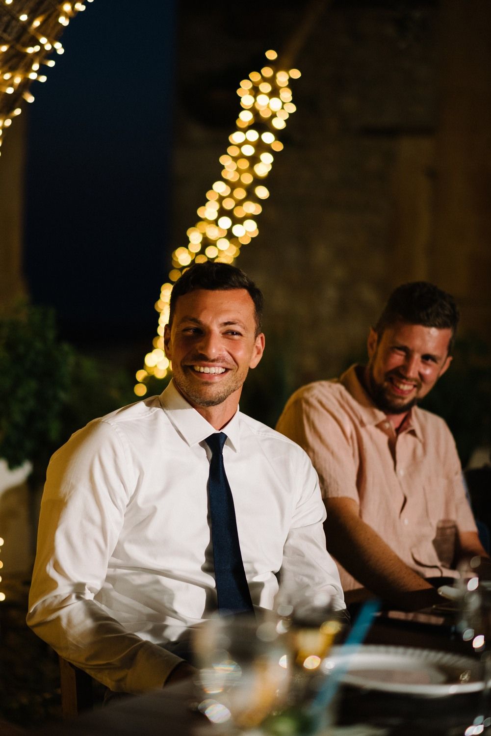 Guests seated at an outdoor dinner table beneath trees illuminated with string lights.