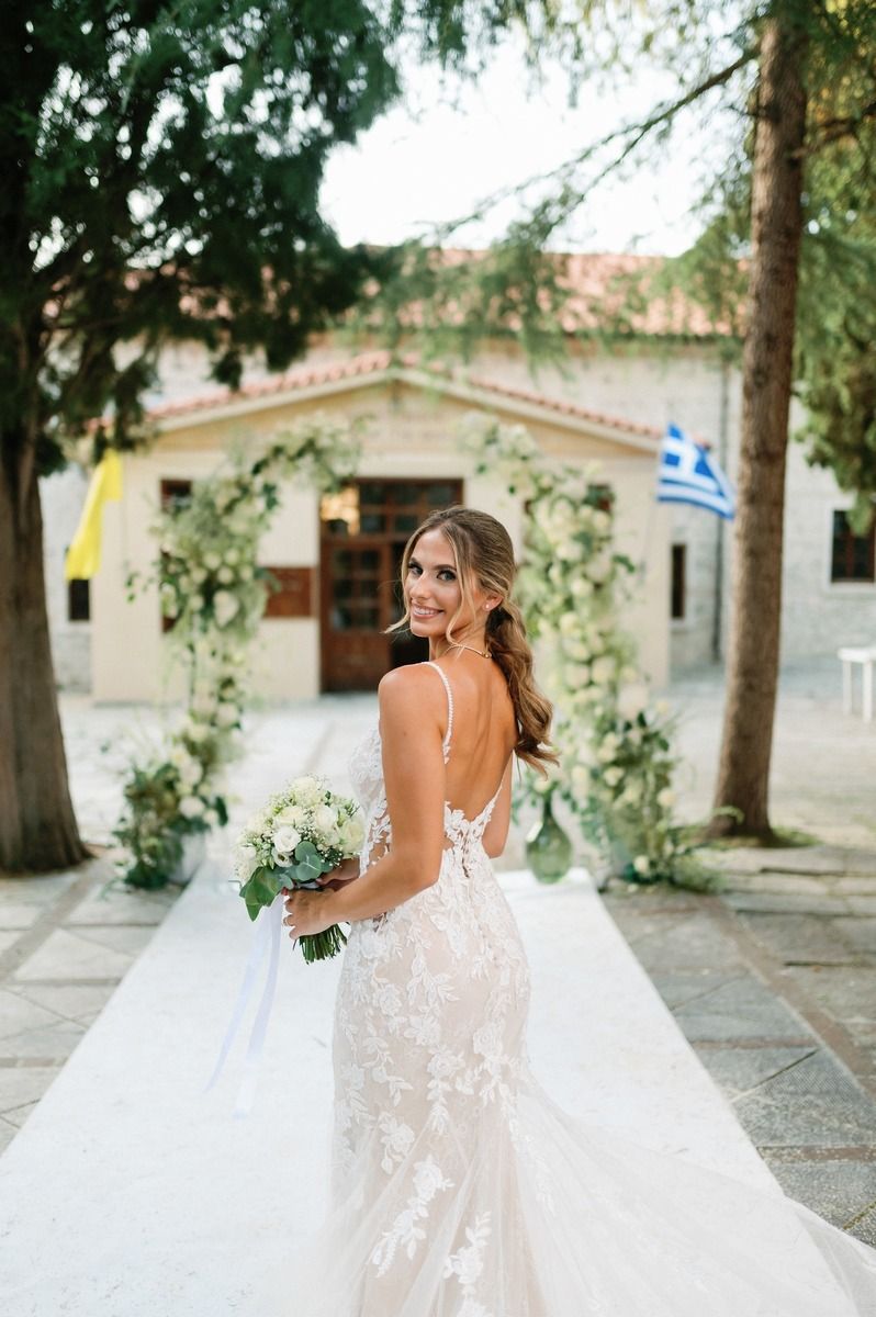 Bride in lace wedding dress holds bouquet against Mediterranean architecture.