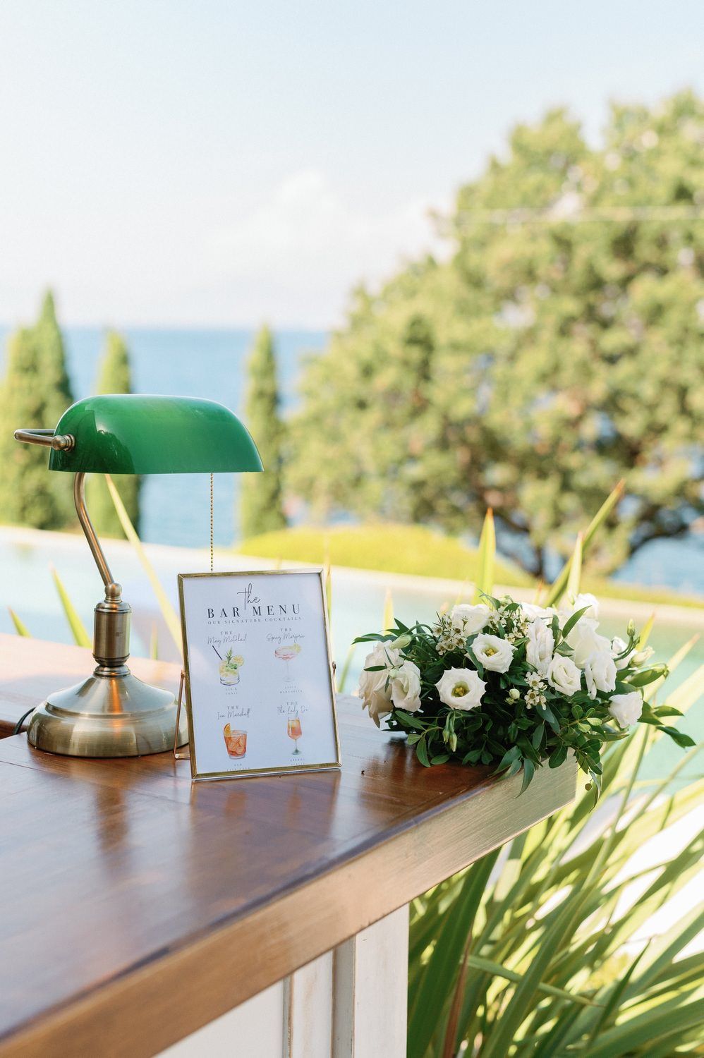 Bar menu setup with green banker's lamp and white floral arrangement on wooden table.