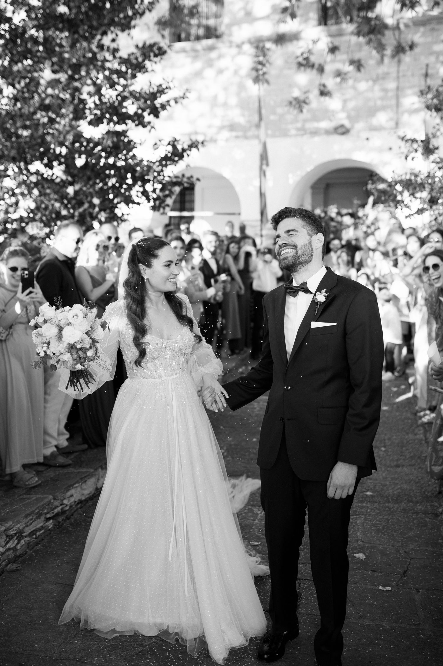 A bride in a flowing white gown and groom in a tuxedo share a joyful moment during their outdoor wedding celebration.