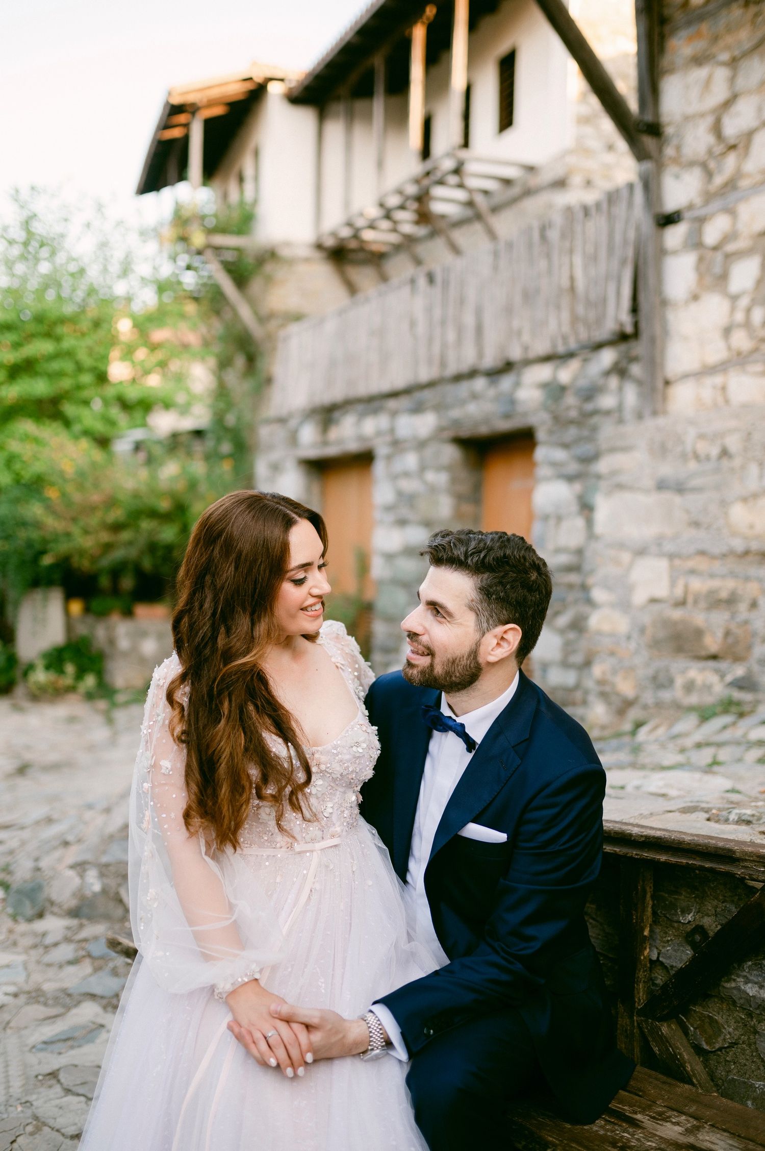 Elegant couple in wedding attire sitting together on stone steps by rustic building with romantic ambiance.