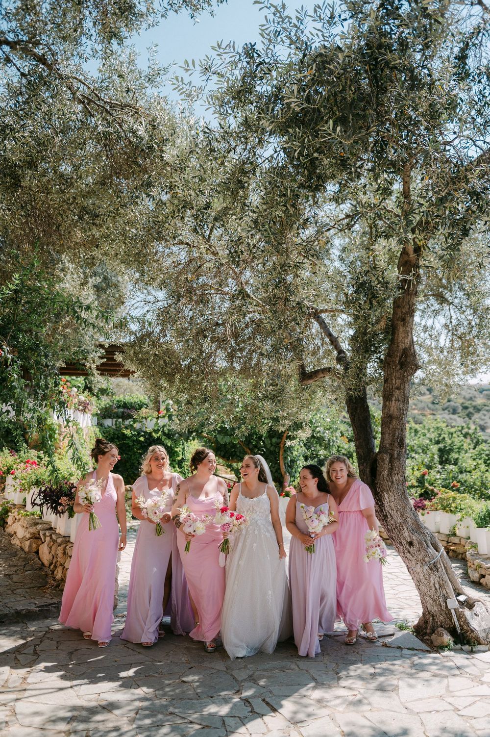 A group in pink bridesmaid dresses pose together under olive trees in a sun-dappled Mediterranean garden setting.