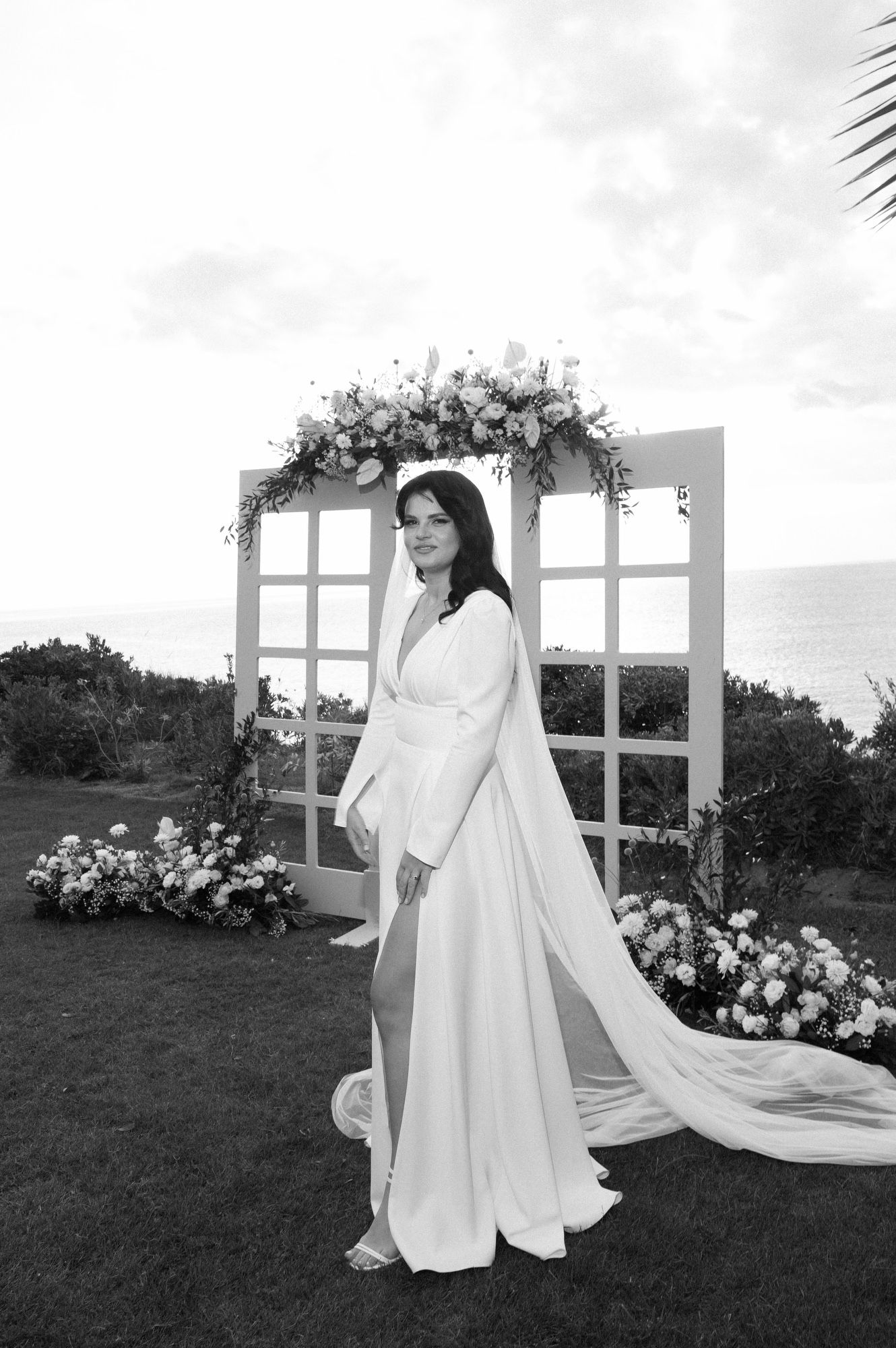 Elegant bride in a modern white wedding gown with long flowing cape poses in front of scenic outdoor venue.