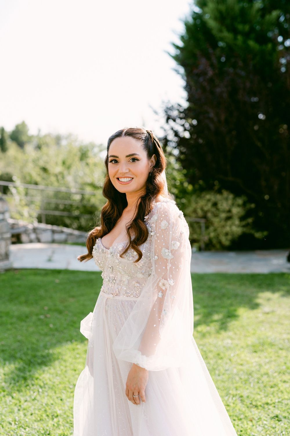 A person in a flowing white lace dress poses outdoors in a sunlit garden setting.