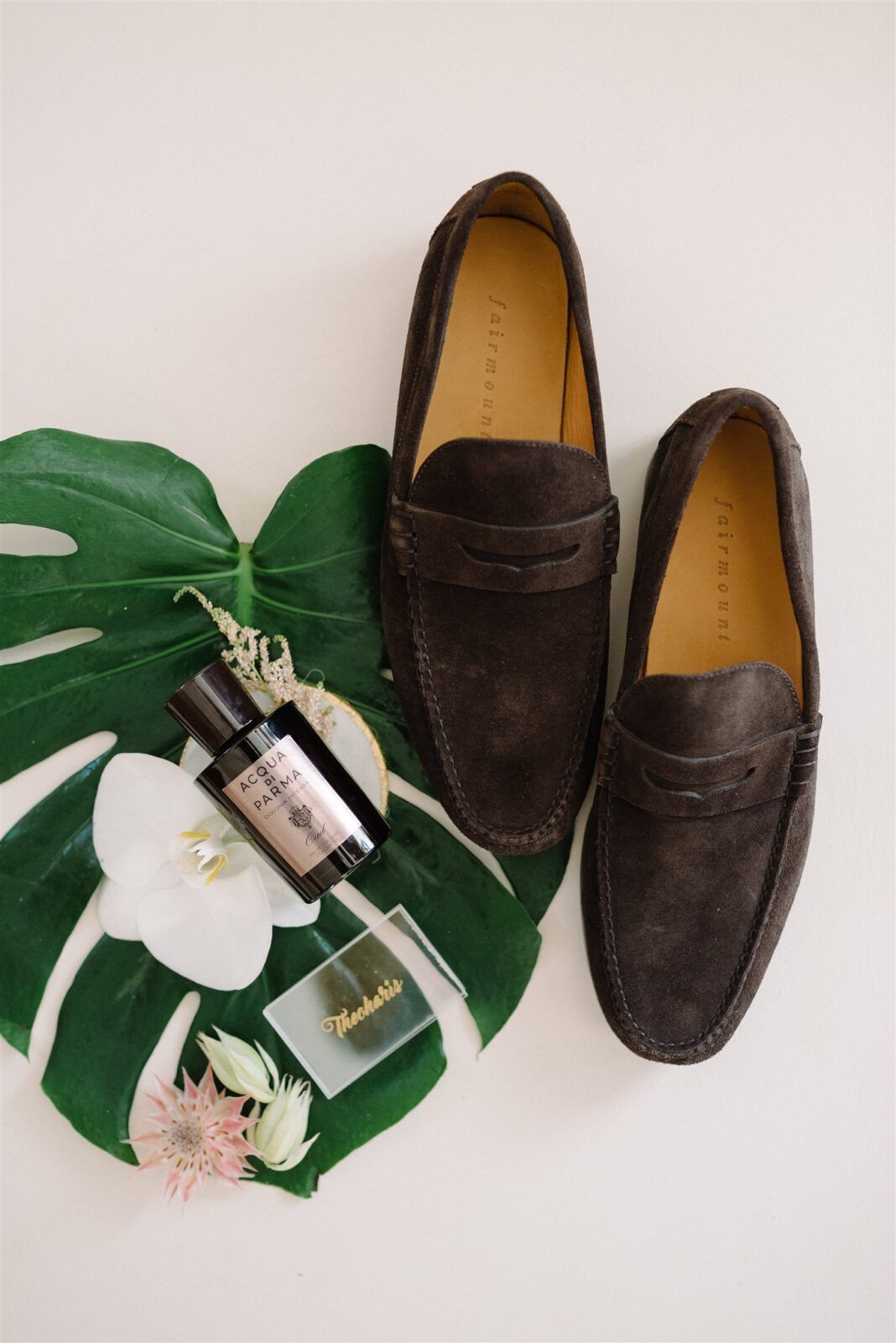 Brown suede loafers styled with tropical monstera leaves and perfume bottles in a minimalist flatlay composition.