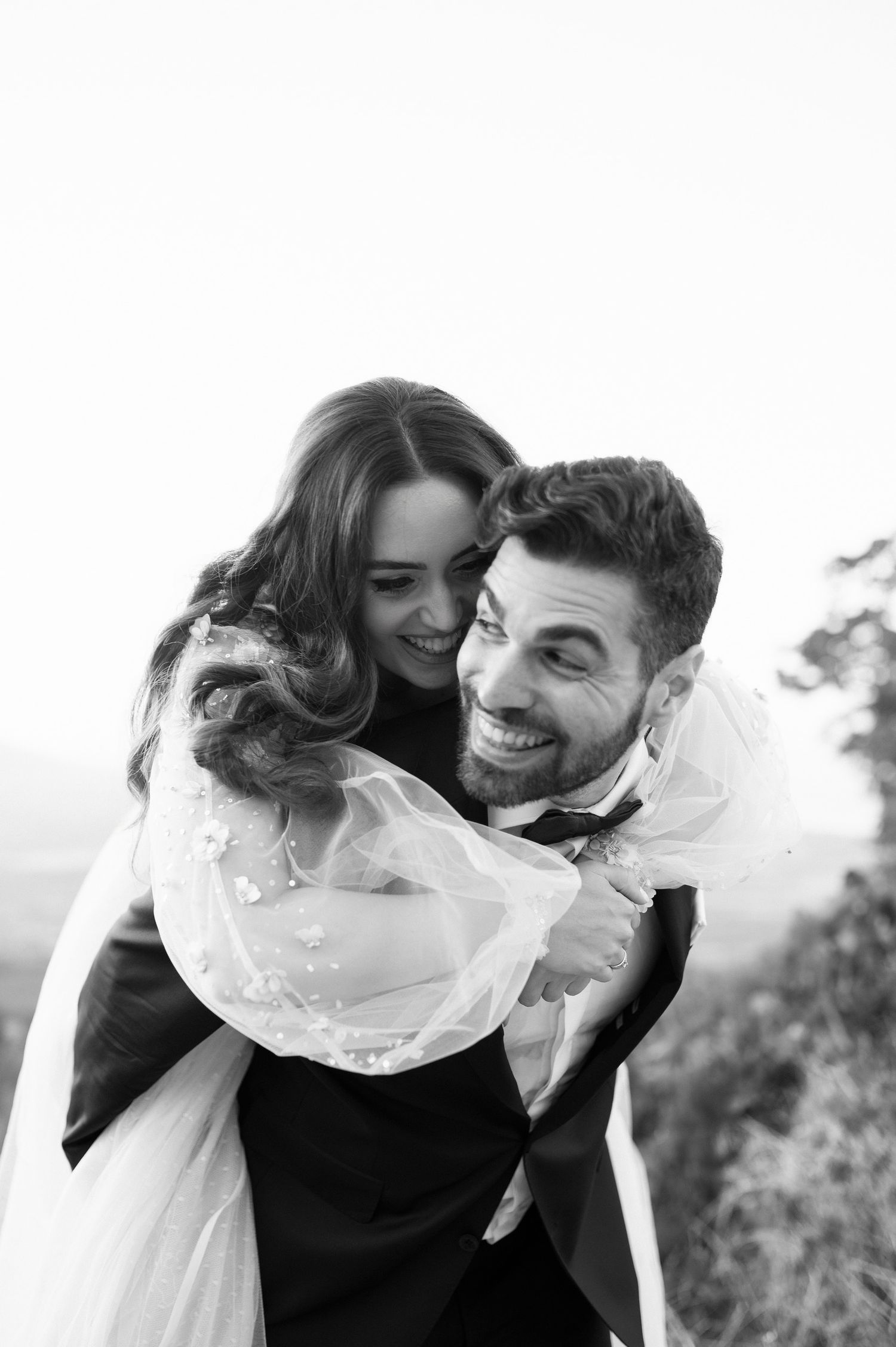 A joyful couple shares an intimate moment in black and white as the bride rides piggyback on the groom outdoors.