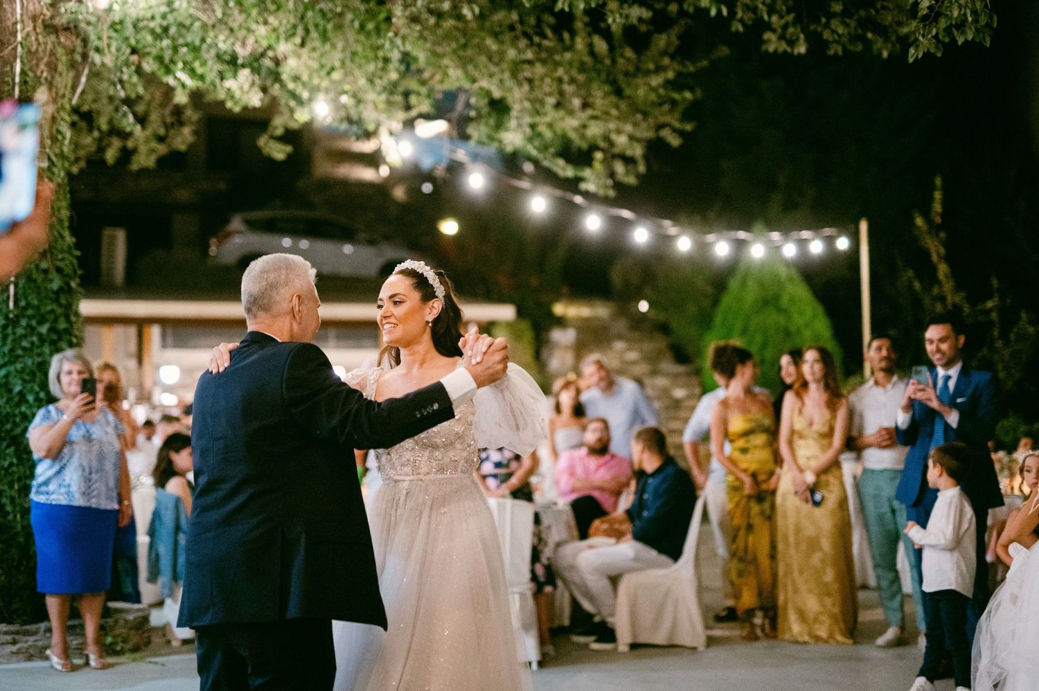 Two people share a dance under outdoor string lights at an evening wedding reception while guests look on.