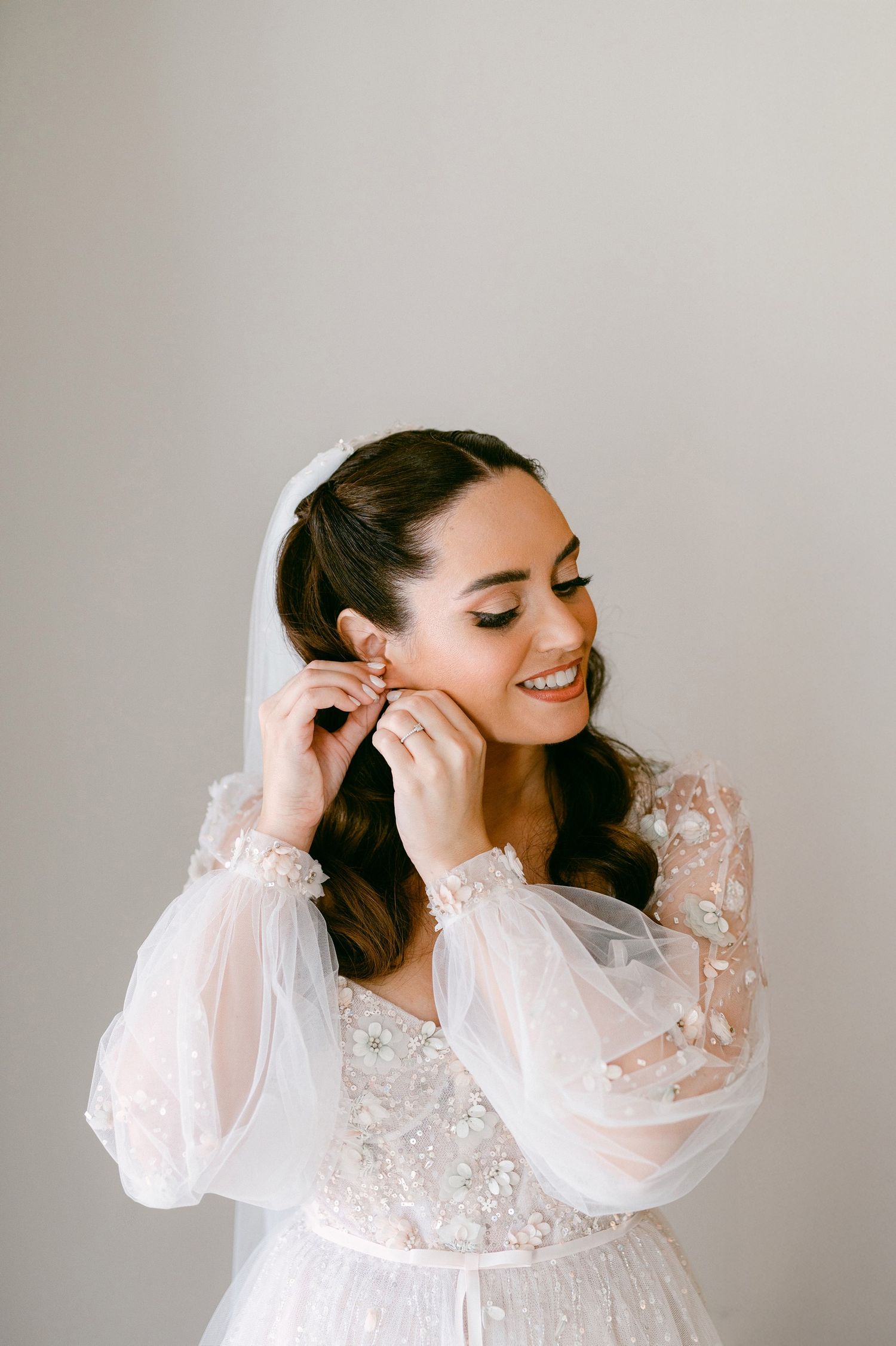 A bride wearing a delicate white lace dress with puff sleeves puts on earrings while smiling with eyes downcast.