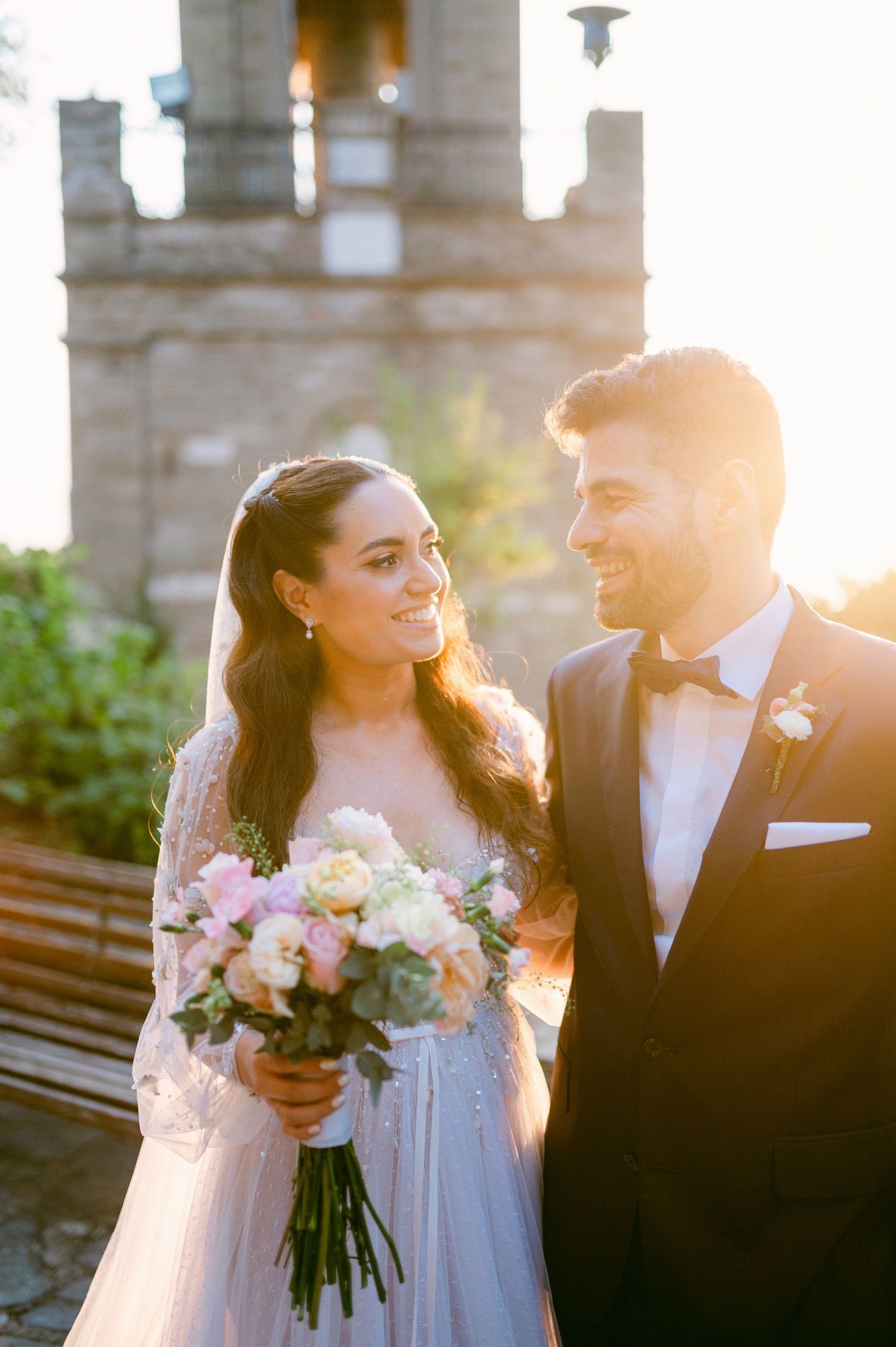 A romantic wedding moment captured at sunset with a bride holding a blush and white bouquet next to a groom in a tuxedo.