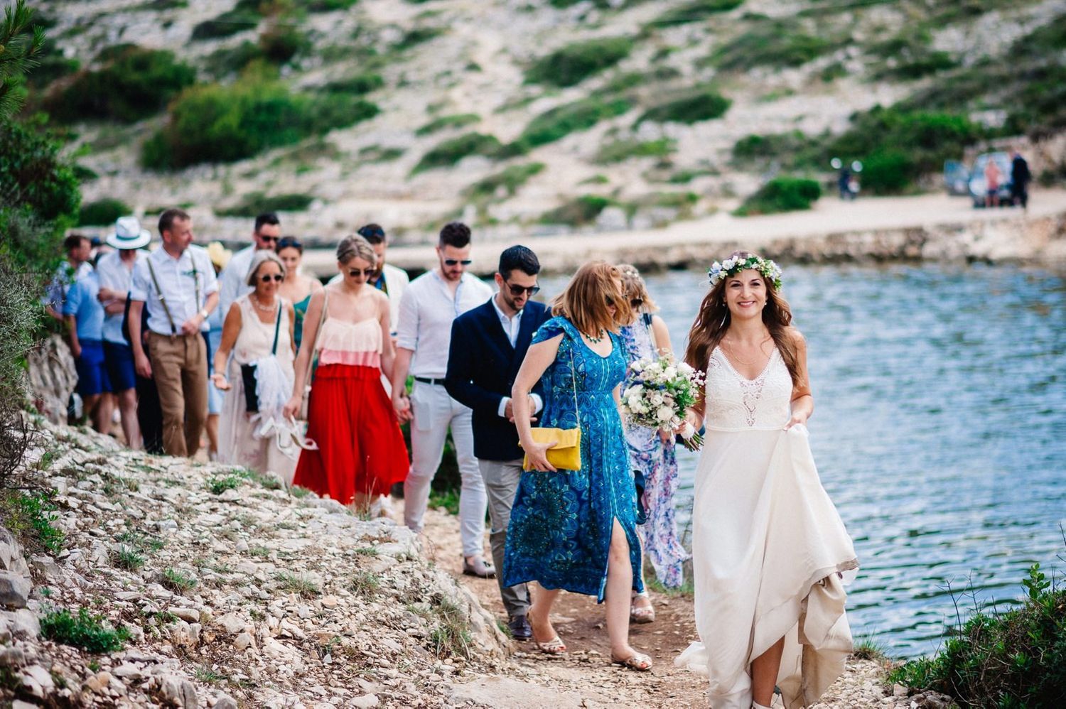 Wedding party walks along rocky Mediterranean coastline during seaside celebration.