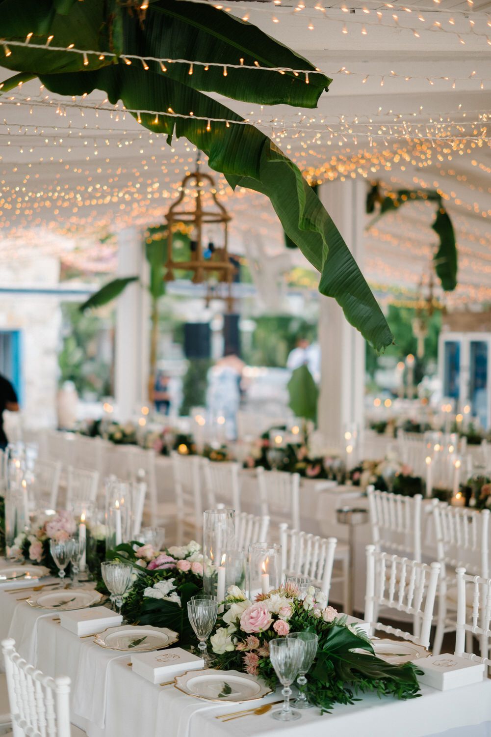 Tropical wedding reception decor with string lights, banana leaves, and white table settings.