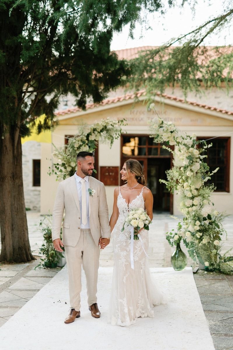 Elegant wedding couple stands on white carpet in front of Mediterranean style building with floral arch.