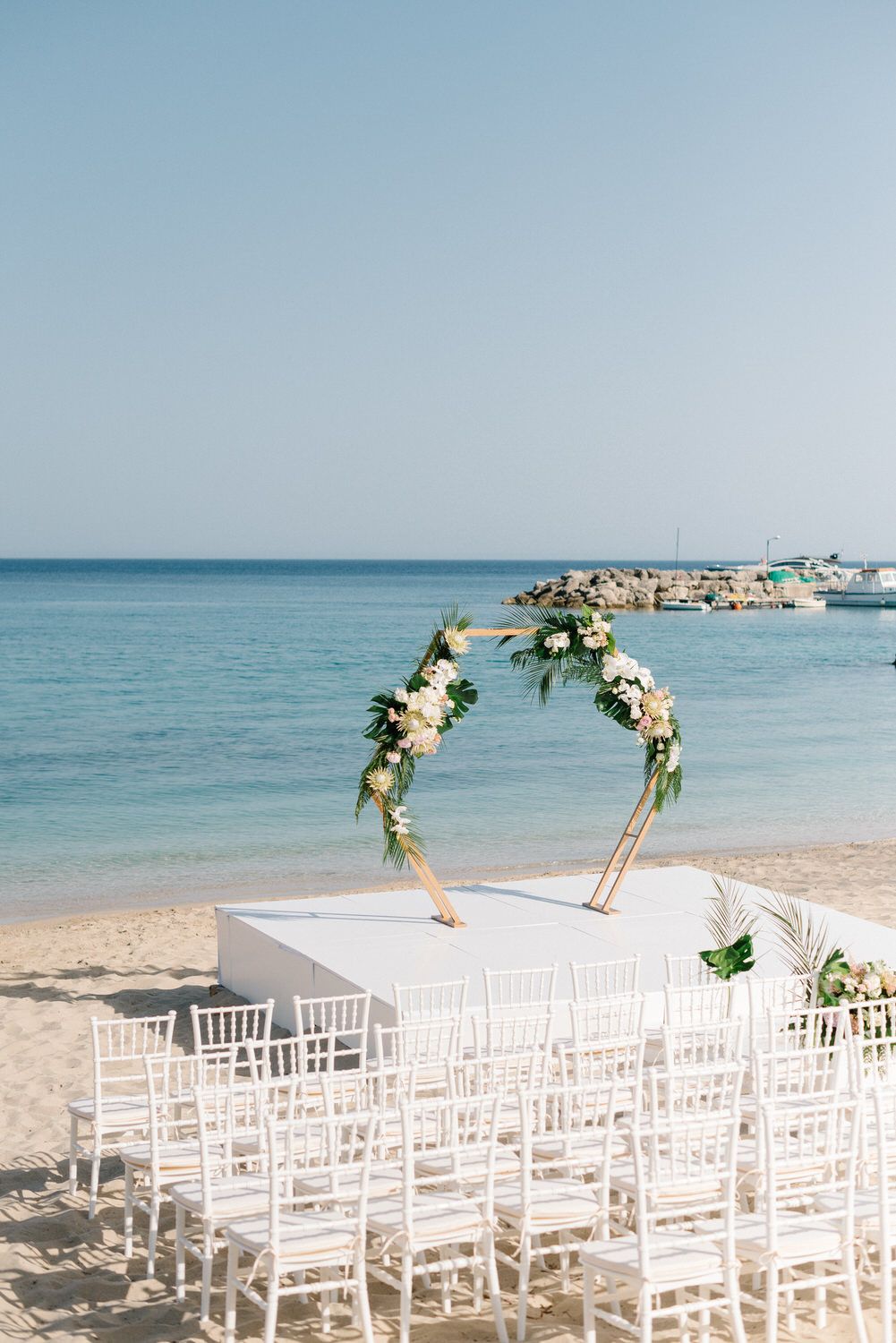 White chairs set up for beach wedding ceremony with floral arch and ocean backdrop.
