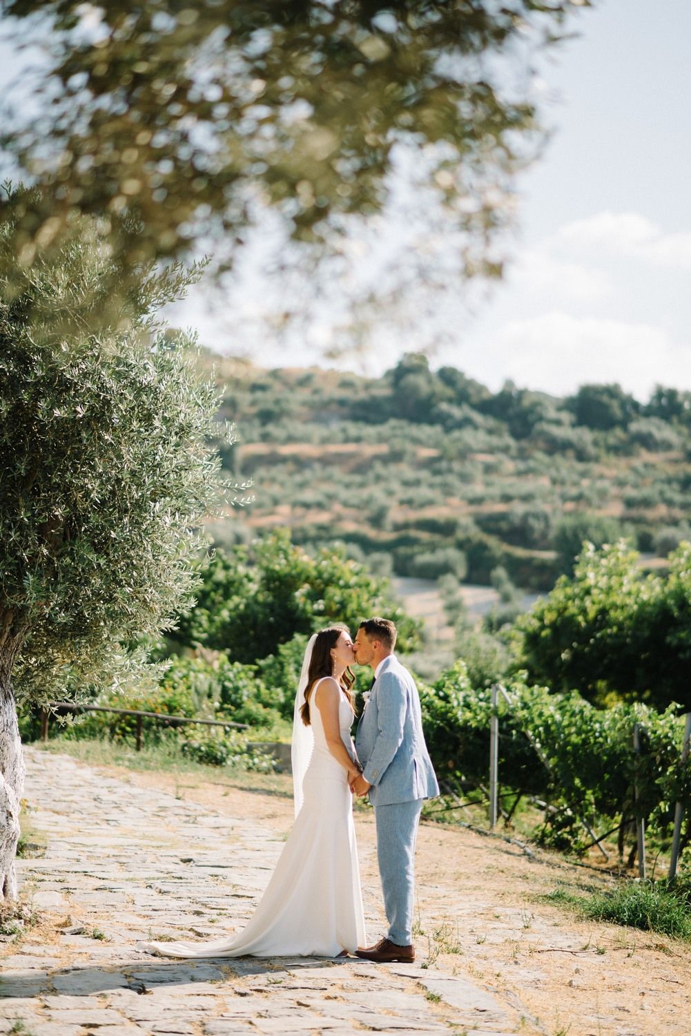 Bride and groom share an intimate moment on a stone path surrounded by vineyard and olive trees.