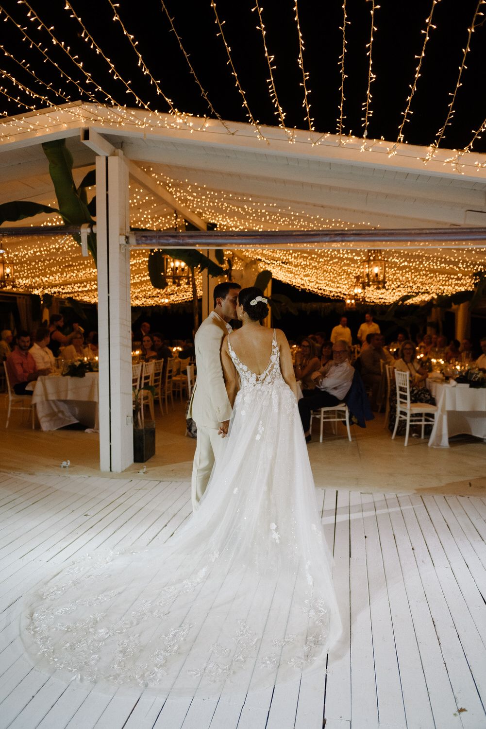 Stunning bridal gown with long train photographed under a ceiling of glowing string lights.