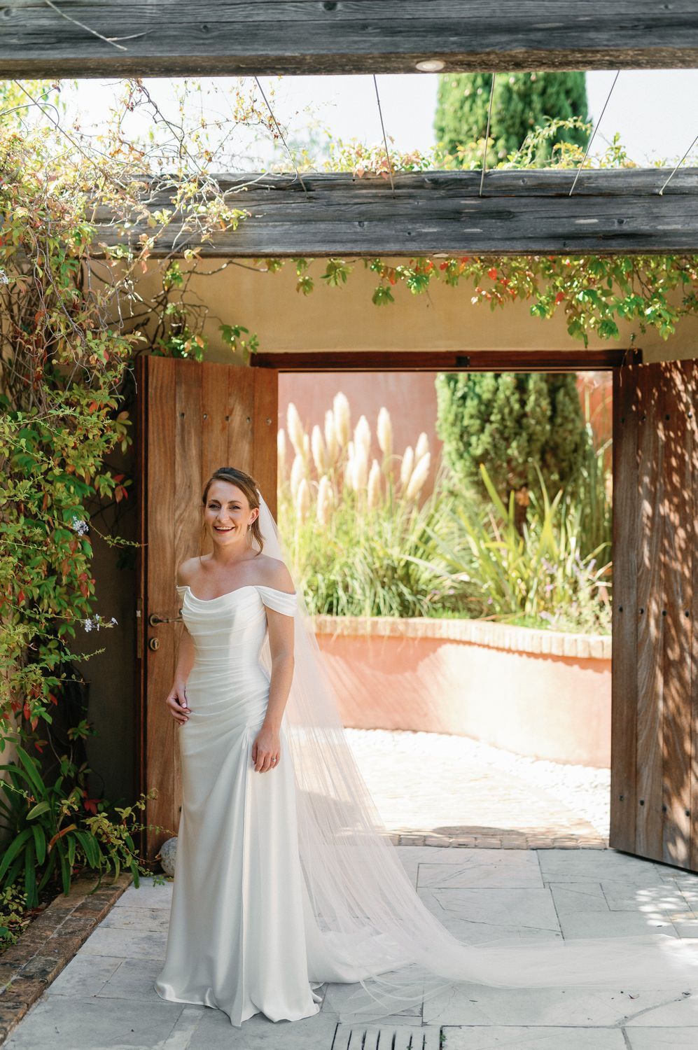 A bride in an elegant off-shoulder gown stands under a rustic wooden pergola with trailing vines.